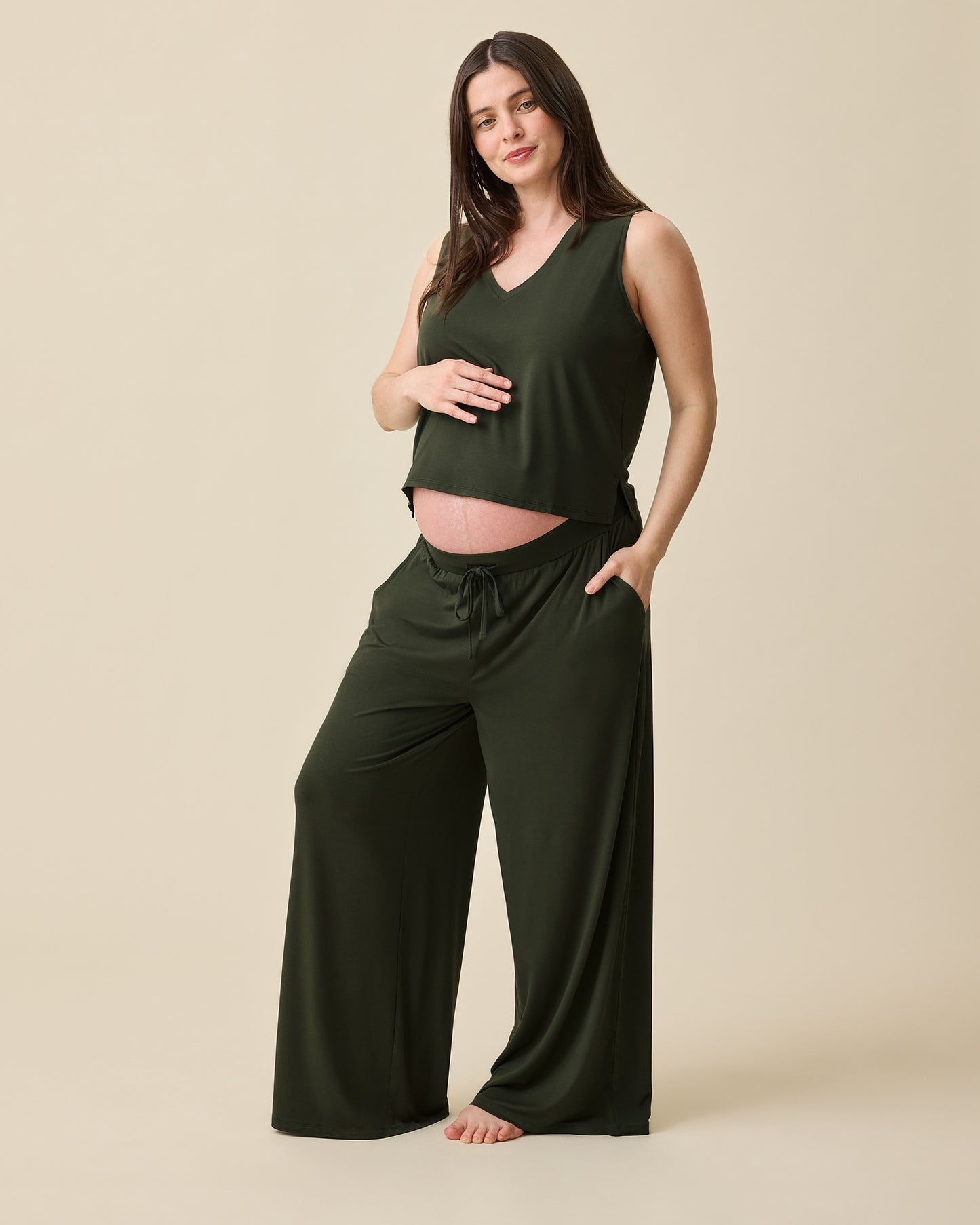 Model is pregnant and wearing Aria Bamboo Cropped V-neck Tank in Thyme and Aria Bamboo Casual Lounge Pant in Thyme on a beige background.@model_info:Tassia is 5'6" and wearing a Medium.