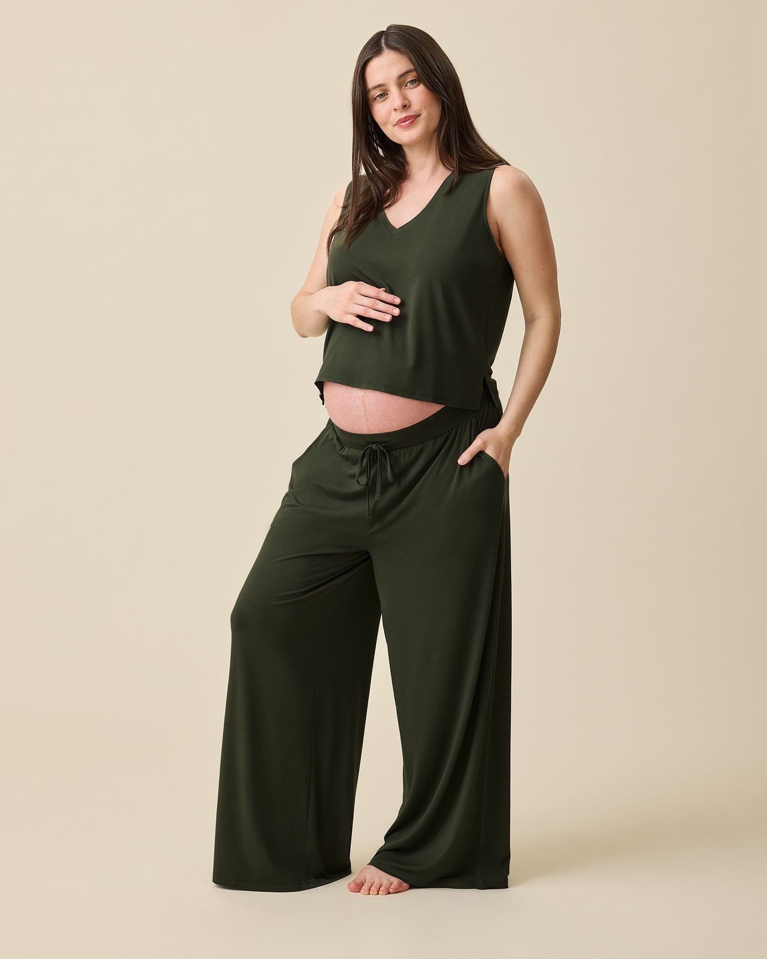 Model is pregnant and wearing Aria Bamboo Cropped V-neck Tank in Thyme and Aria Bamboo Casual Lounge Pant in Thyme on a beige background.@model_info:Tassia is 5'6" and wearing a Medium.