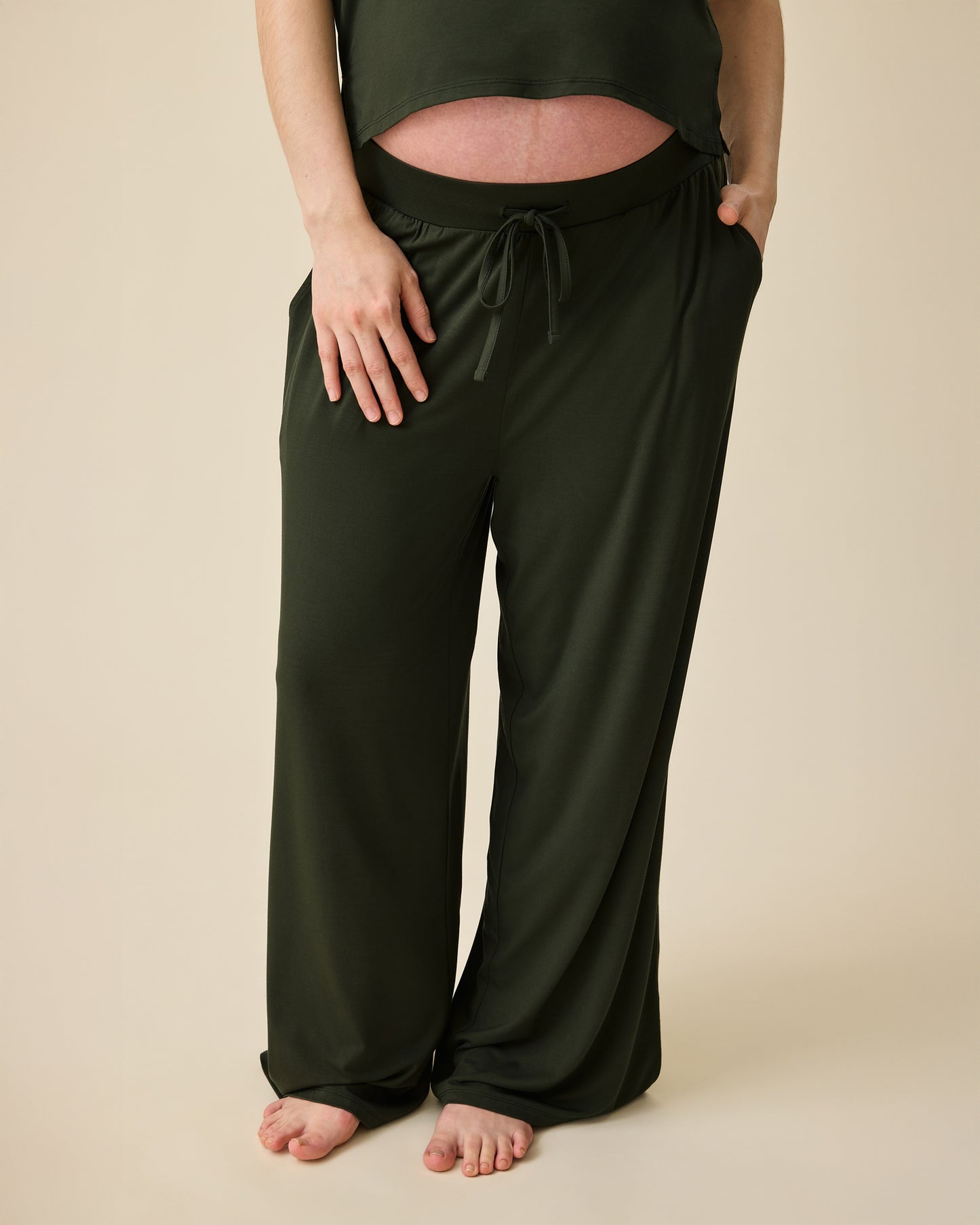 Model is pregnant and wearing Aria Bamboo Cropped V-neck Tank in Thyme and Aria Bamboo Casual Lounge Pant in Thyme on a beige background.