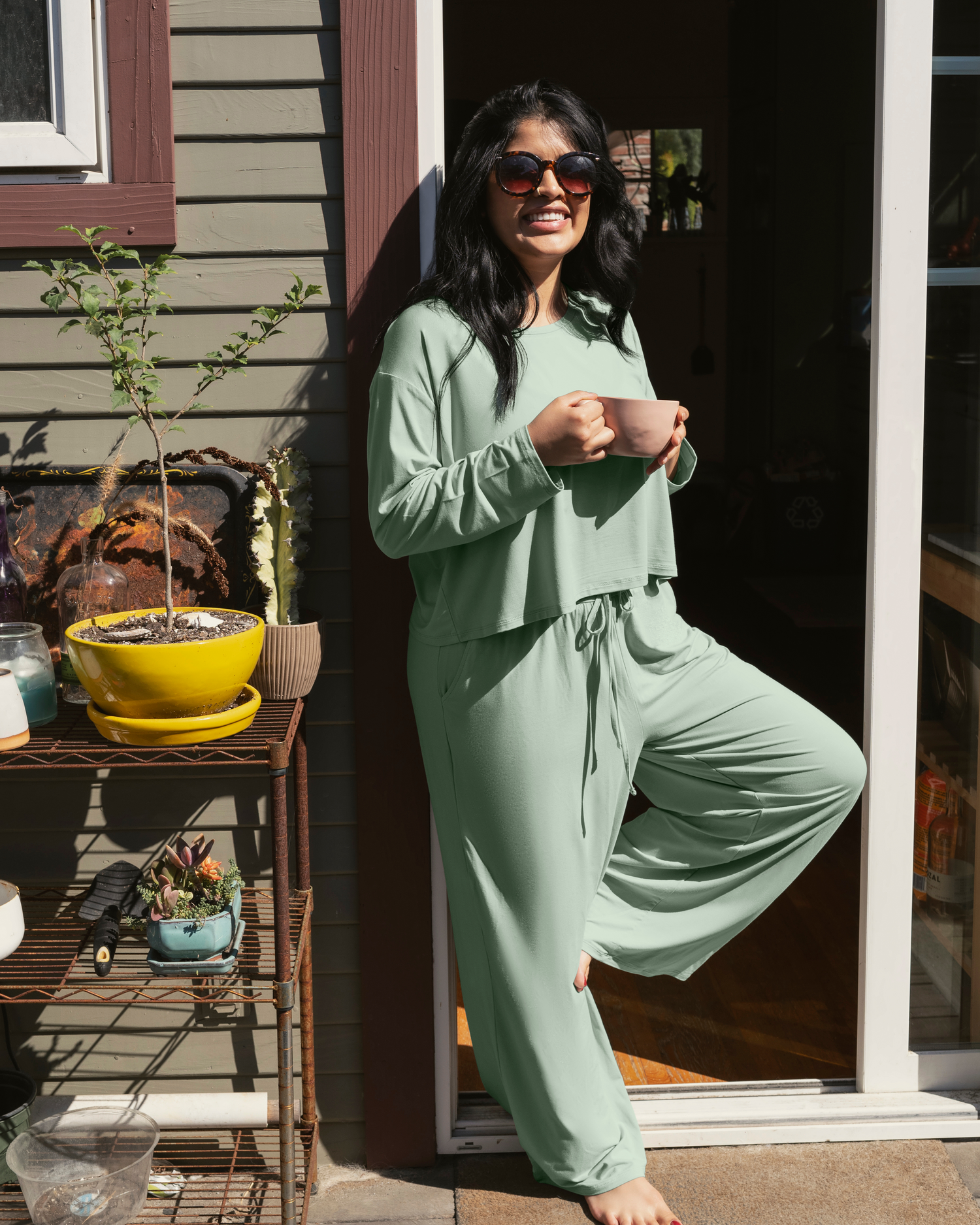 Model outside with coffee mug, leaning against house wearing the Aria Bamboo Casual Lounge Pant in Aqua Mist paired with matching Aria Bamboo Casual Cropped Long Sleeve Top @model_info:Betsabe is 5'5" and wearing a Medium.