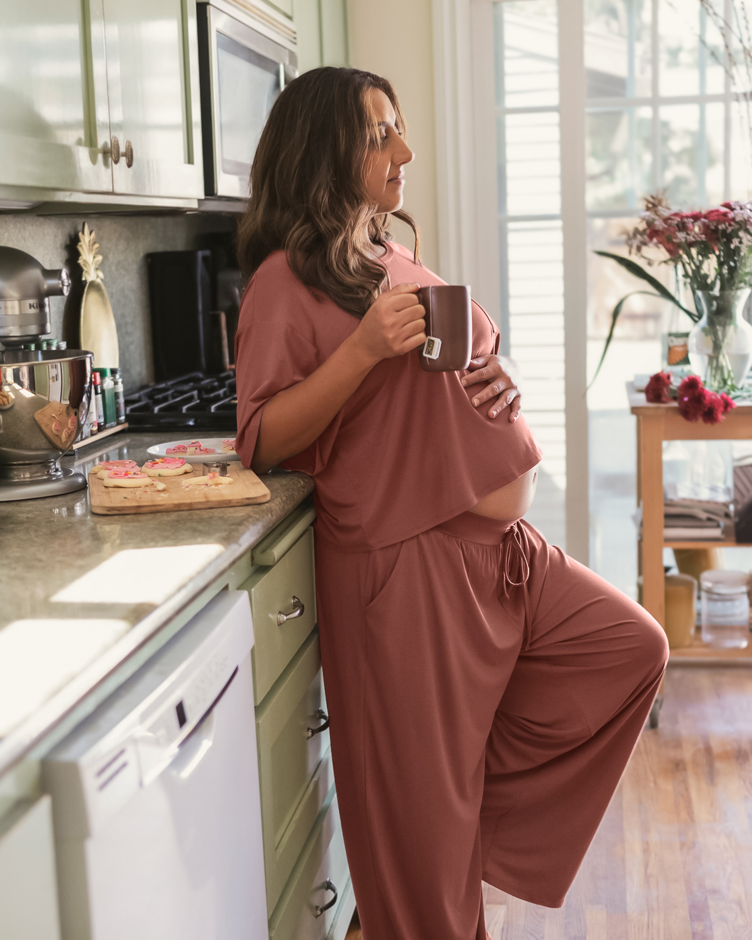 Side view of pregnant model wearing the  Aria Bamboo Casual Lounge Pant in Redwood, paired with matching Aria Bamboo Casual Cropped Short Sleeve Top, leaning against kitchen counter and holding mug of tea @model_info:Jaqueline is 5'3" and wearing a Large.