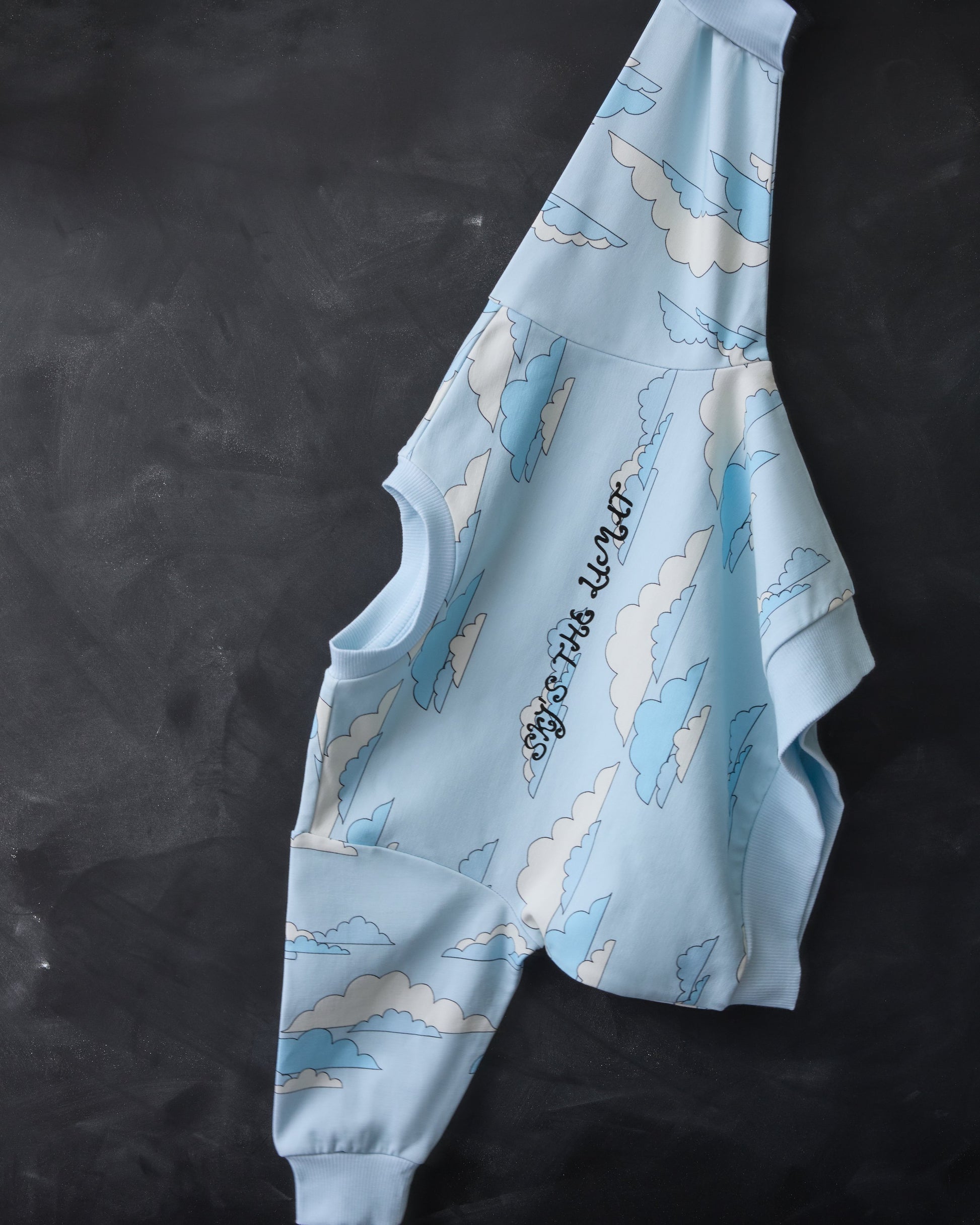 Graphic Sweatshirt in Blue Clouds is hanging by a sleeve, across a black chalkboard background.