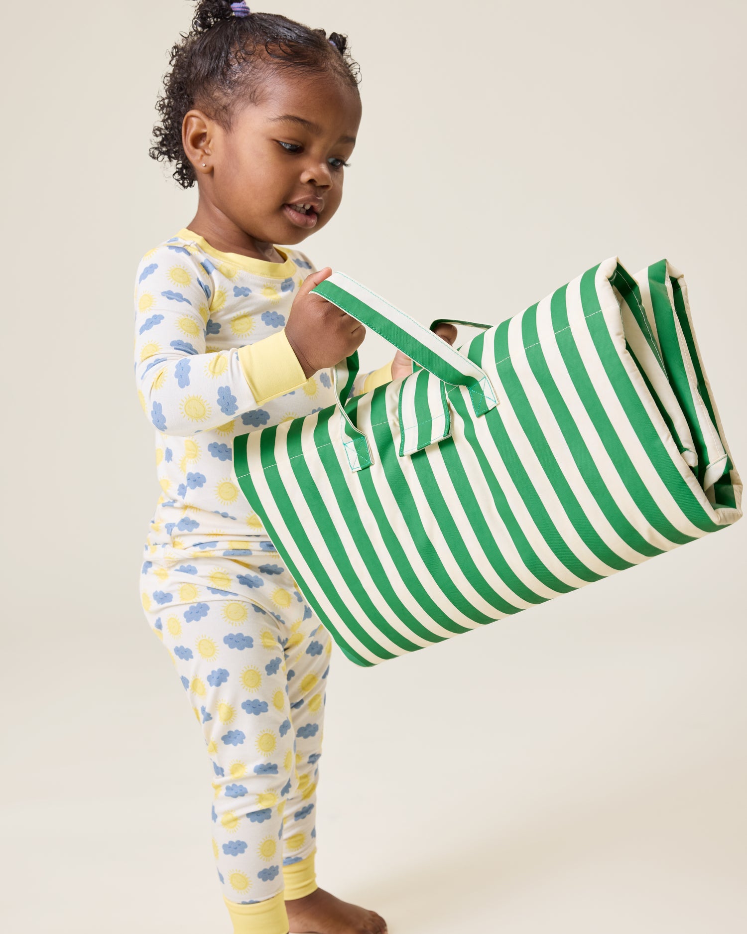Child in Sunny Day print pajamas holding the The Inside/Outside Play Mat in Green Stripe print folded into a bag