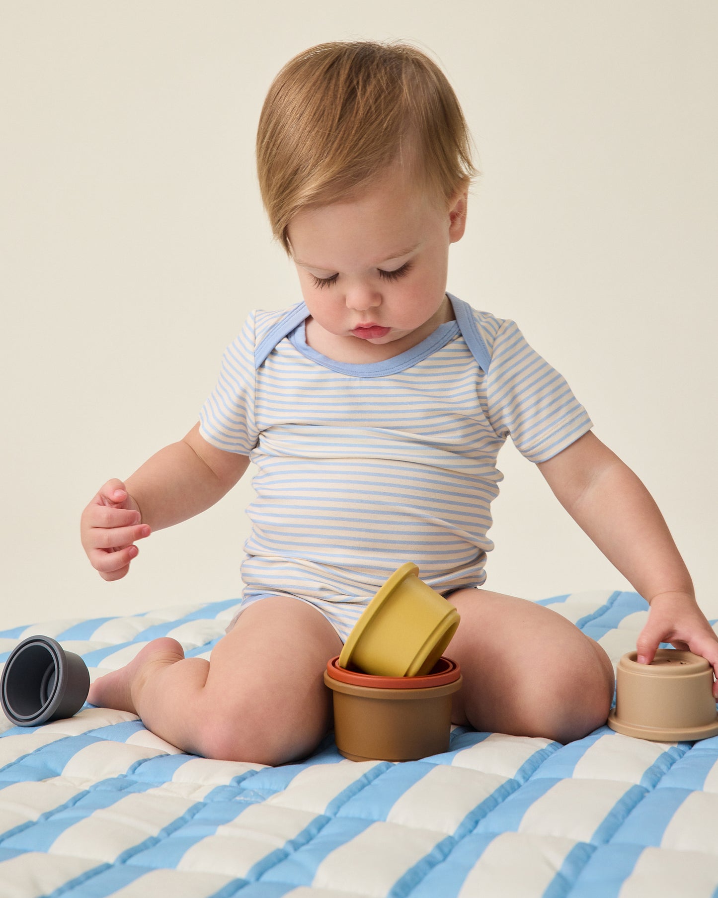Baby playing with stack and nest cup toys on Inside/Outside Play Mat in Blue Wide Stripe, on a beige background