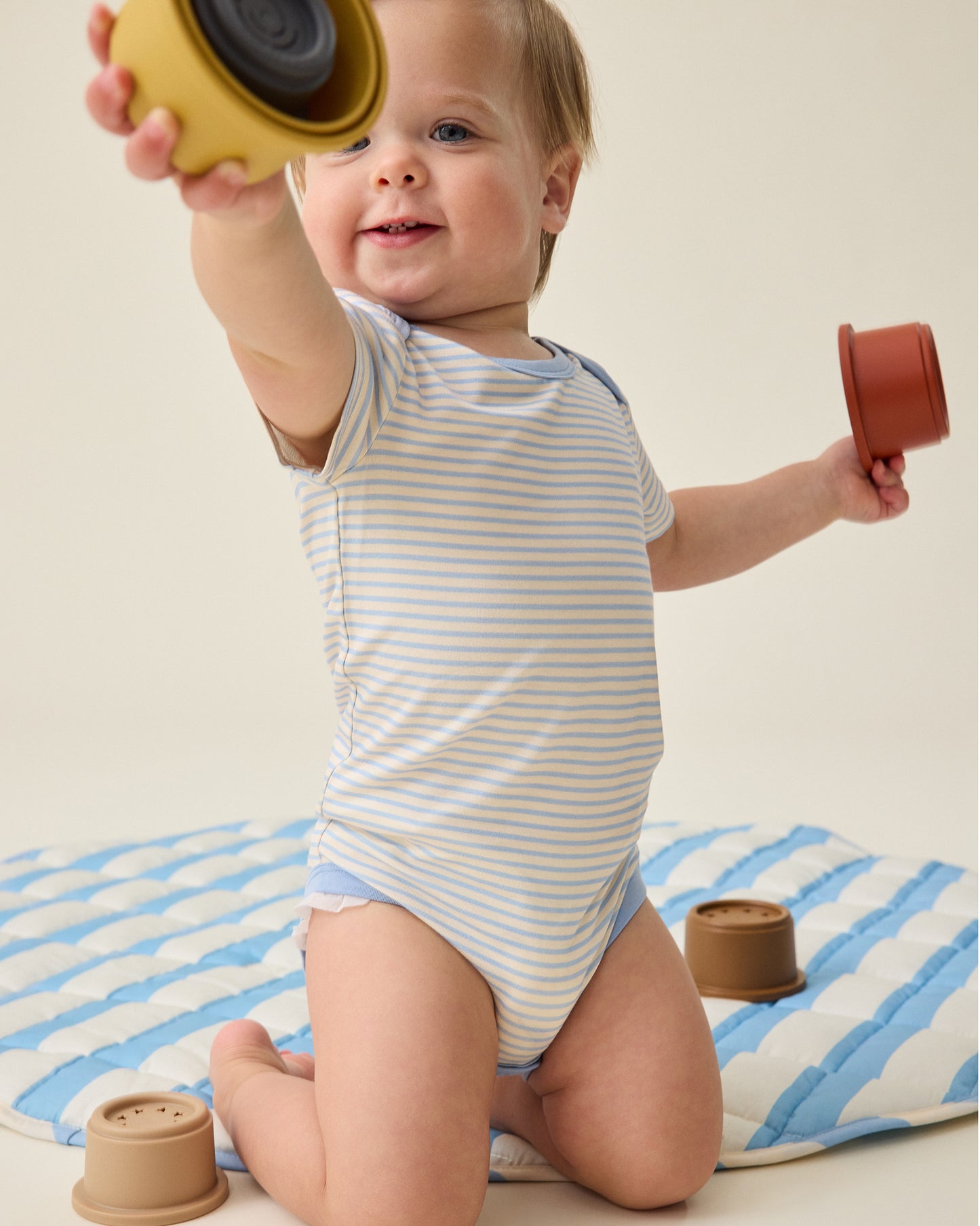 Baby wearing a blue and white striped onesie on play mat and playing with toys
