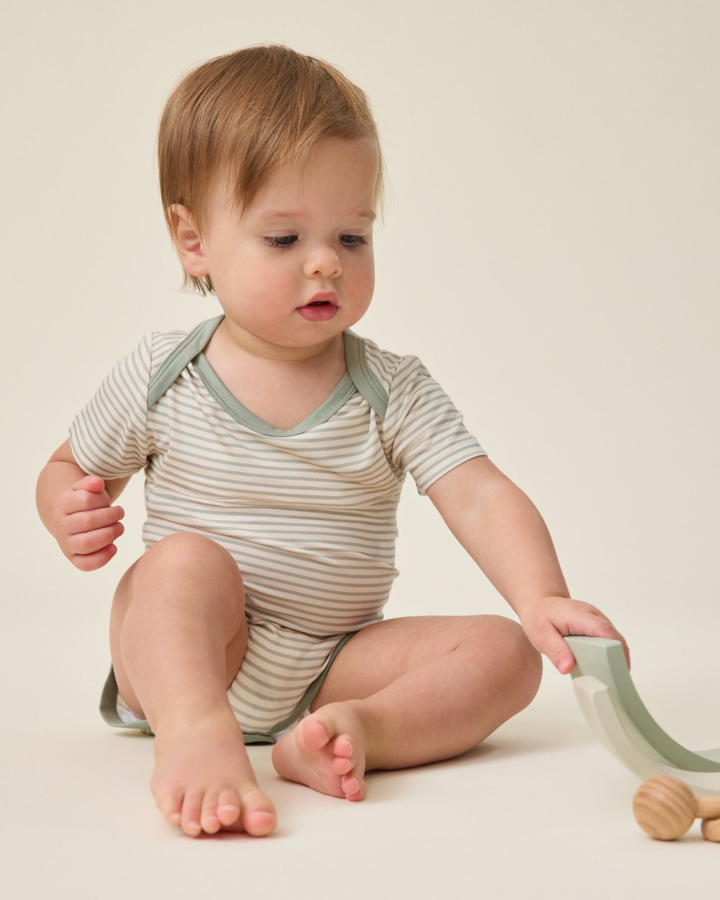 Baby in a striped sage onesie climbing a small green step on a plain background@model_info:Noa is wearing 6-12 months.