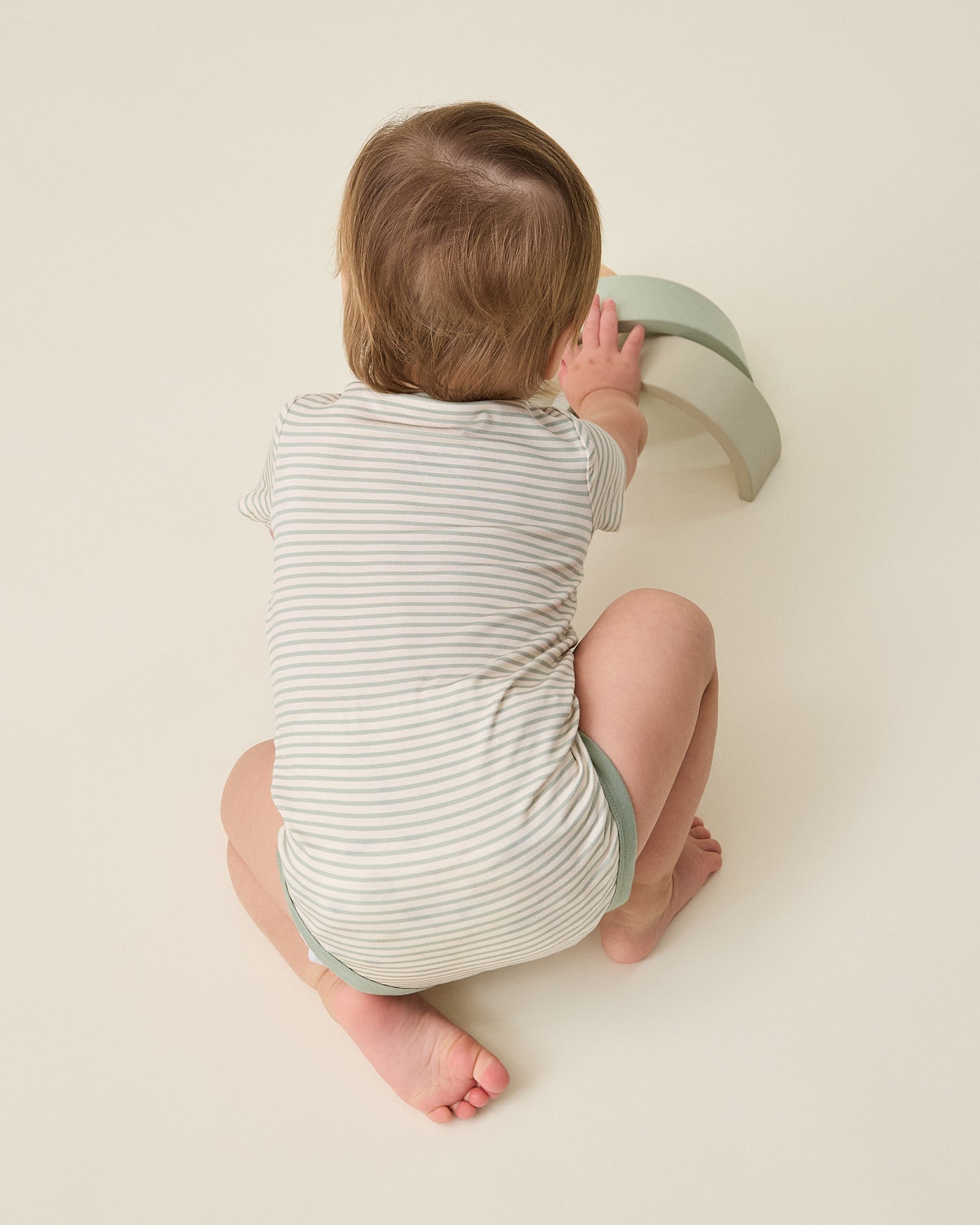 Baby in a striped sage onesie climbing a small green step on a plain background