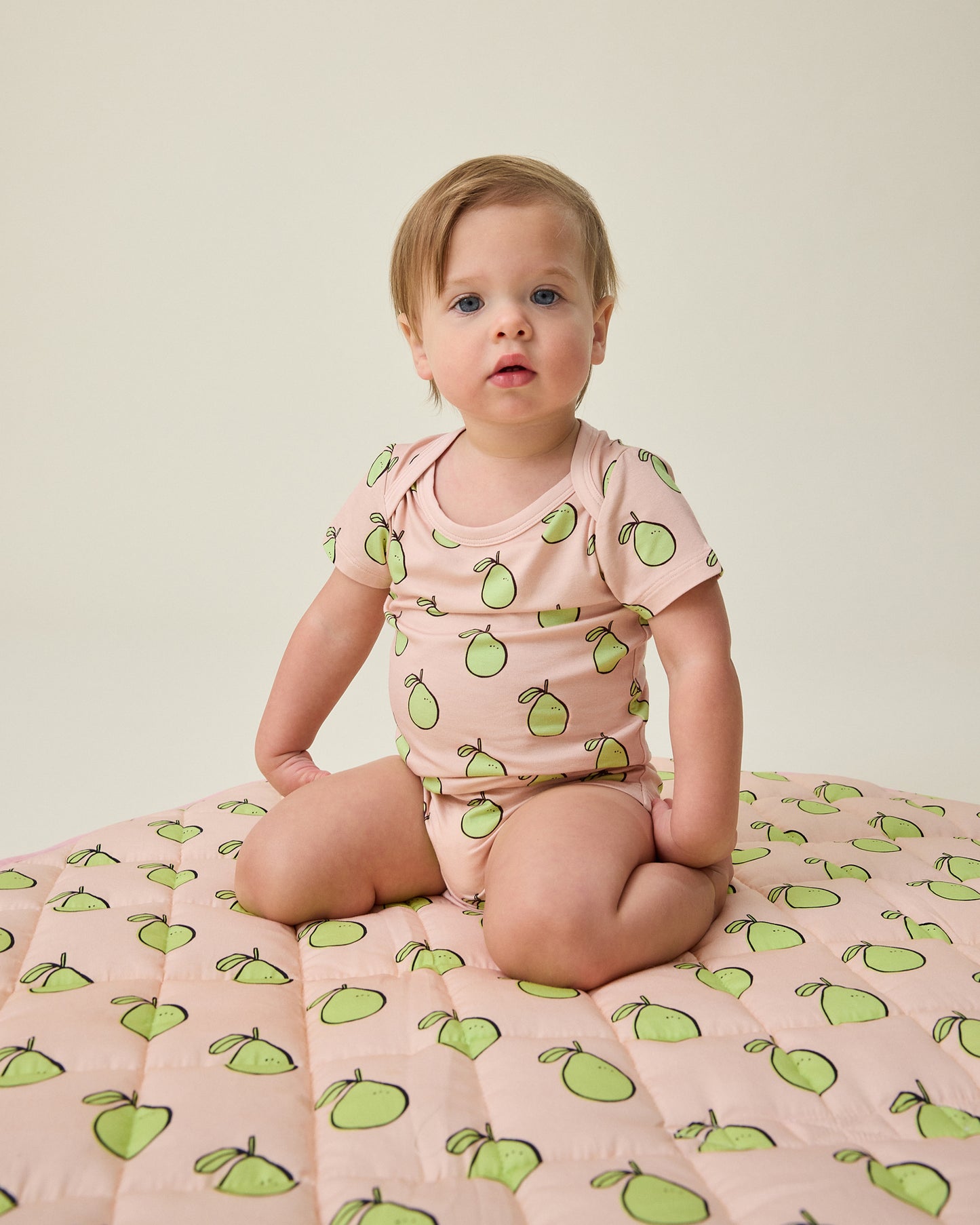 Baby in Pink Pomelo pattern onesie, sitting on Inside/Outside Play Mat in Pink Pomelo pattern, on a beige background