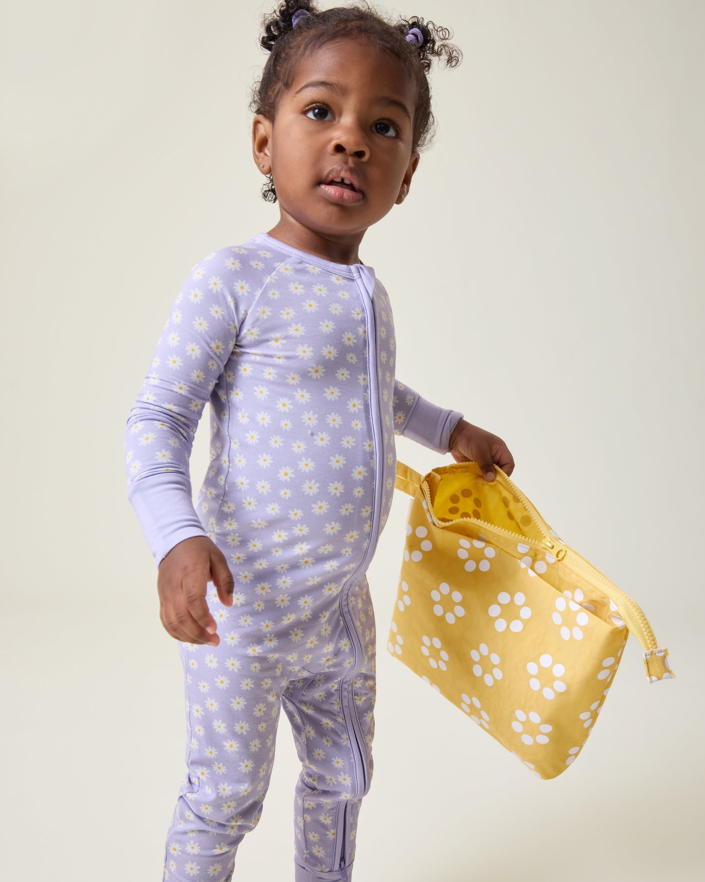 Child wearing Purple Daisies pajamas and holding the Quick Change Kit Bag in Sunshine Dots print, on a neutral background