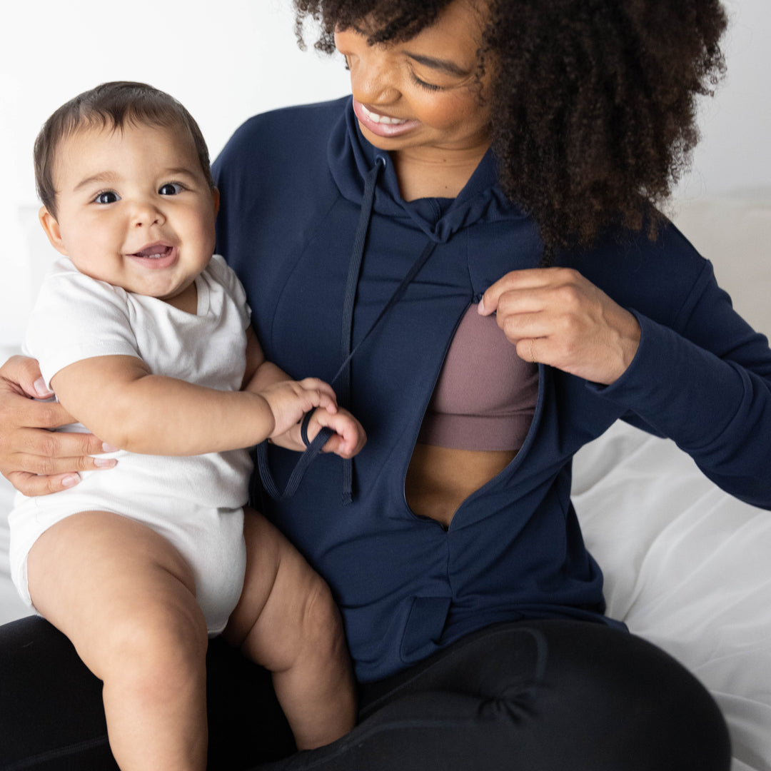 Model wearing the Bamboo Nursing Hoodie in Navy showing the nursing panel and holding her baby on her lap. @Model_info:Tess is wearing a Small.