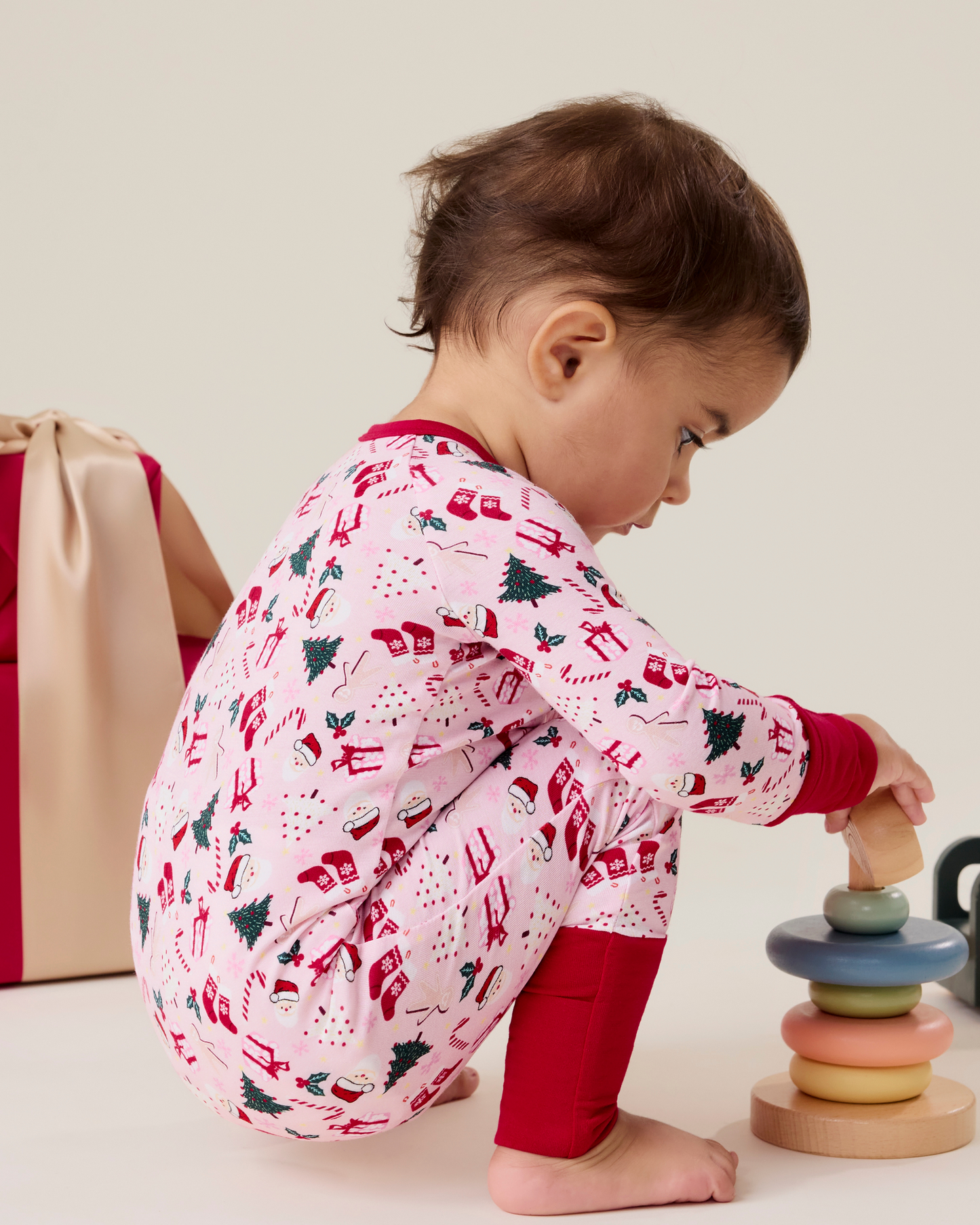 Baby wearing the Bamboo Baby Pajama in Candy Cane Lane - pink with holiday pattern and red trim, playing with toys