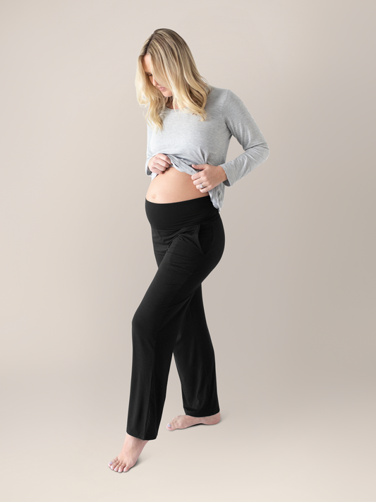Model wearing the Bamboo Maternity & Postpartum Lounge Pant in Black looking down at her belly.@model_info:Maddy is 5'8" and wearing a Medium.