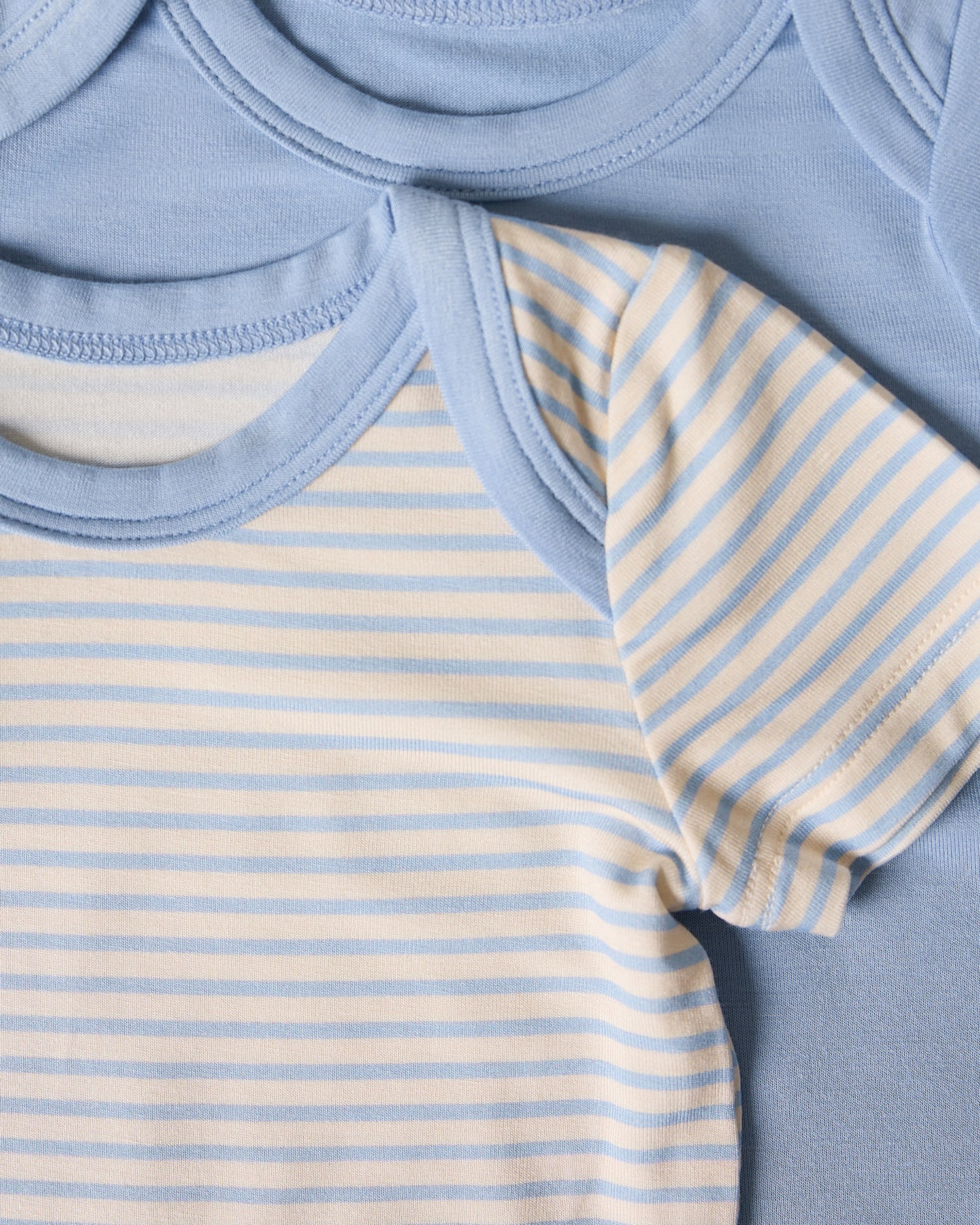 Close-up of two baby bodysuits, one with blue striped pattern and on solid blue on a beige background