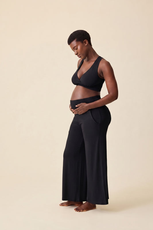 Side view of pregnant model wearing Bamboo Wide Leg Maternity & Postpartum Lounge Pant in Black and French Terry Racerback Nursing & Sleep Bra in Black on a beige background.