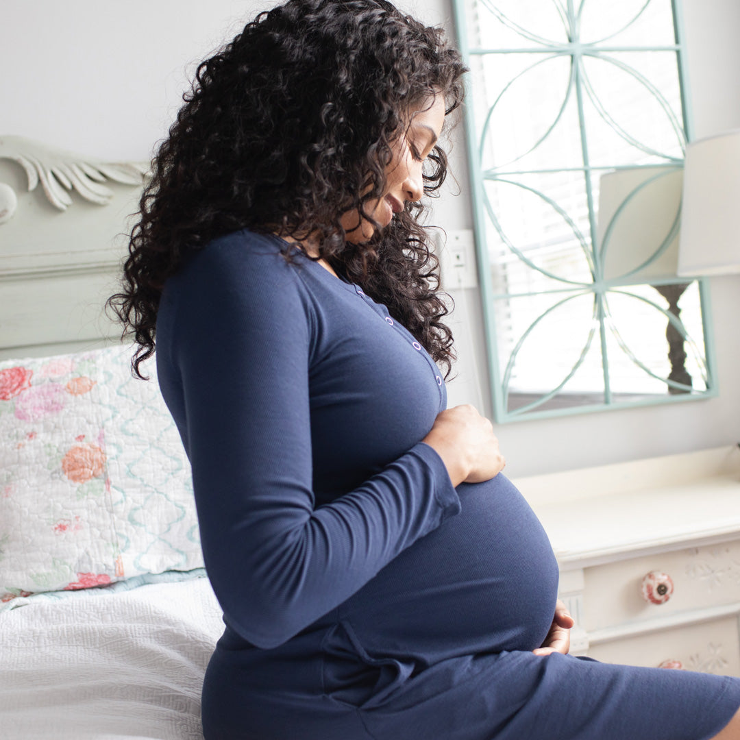 Pregnant model sitting on a bed looking down at her belly and wearing the Betsy Ribbed Bamboo Maternity & Nursing Nightgown