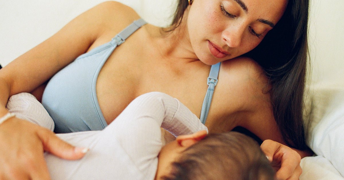 A woman in French Terry Maternity & Nursing Bralette in Vintage Blue is nursing a baby wearing a white long-sleeve top in a softly lit bedroom setting.