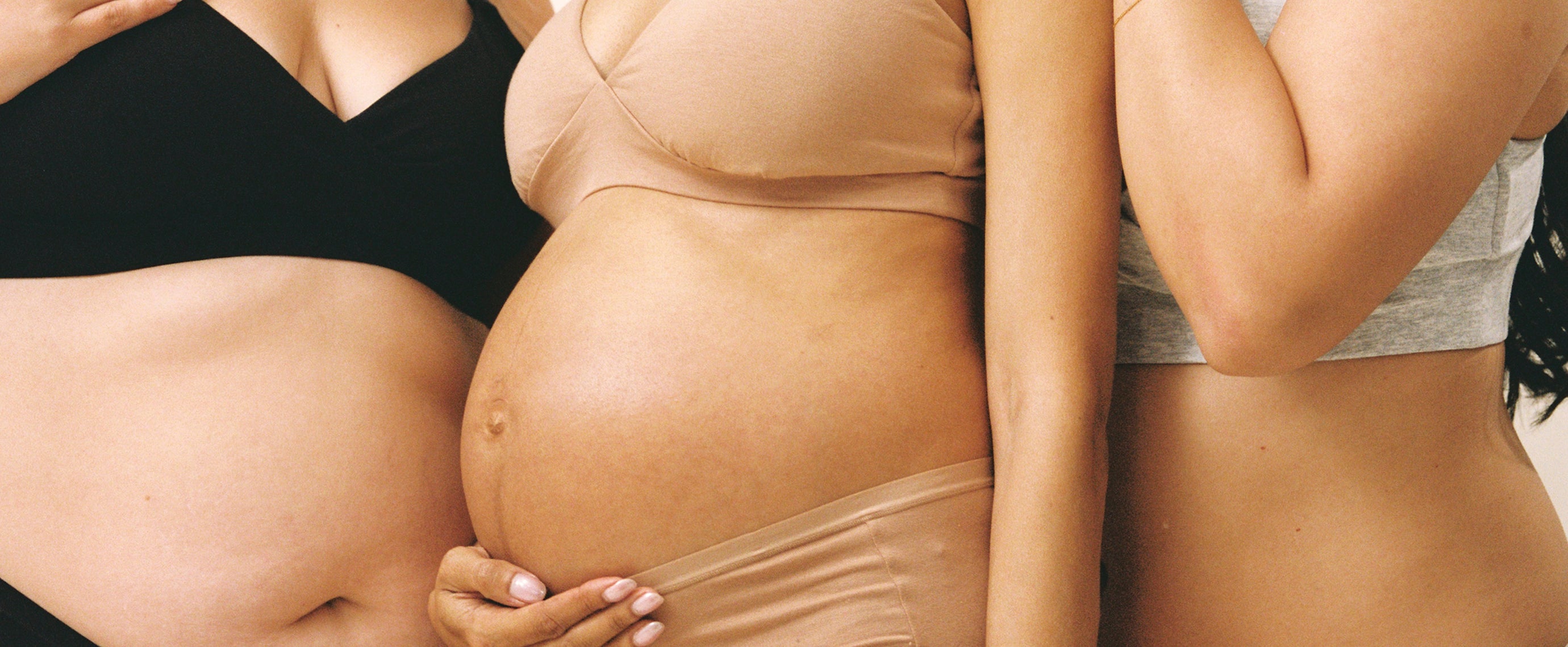 Close up of three models. Postpartum model on the right is wearing SoftStretch Cotton Nursing Bra and SoftStretch Cotton Maternity & Postpartum Hipster in Grey Heather. Pregnant model in the middle is wearing SoftStretch Cotton Maternity & Nursing Bralette and SoftStretch Cotton Maternity & Postpartum Brief in Buff. Model on left is wearing SoftStretch Cotton Nursing Bra and SoftStretch Cotton Maternity & Postpartum Thong in Black on a beige background. 