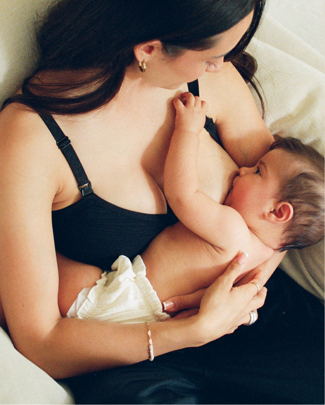 Mother breastfeeding baby while wearing nursing bra, sitting in big white chair. 