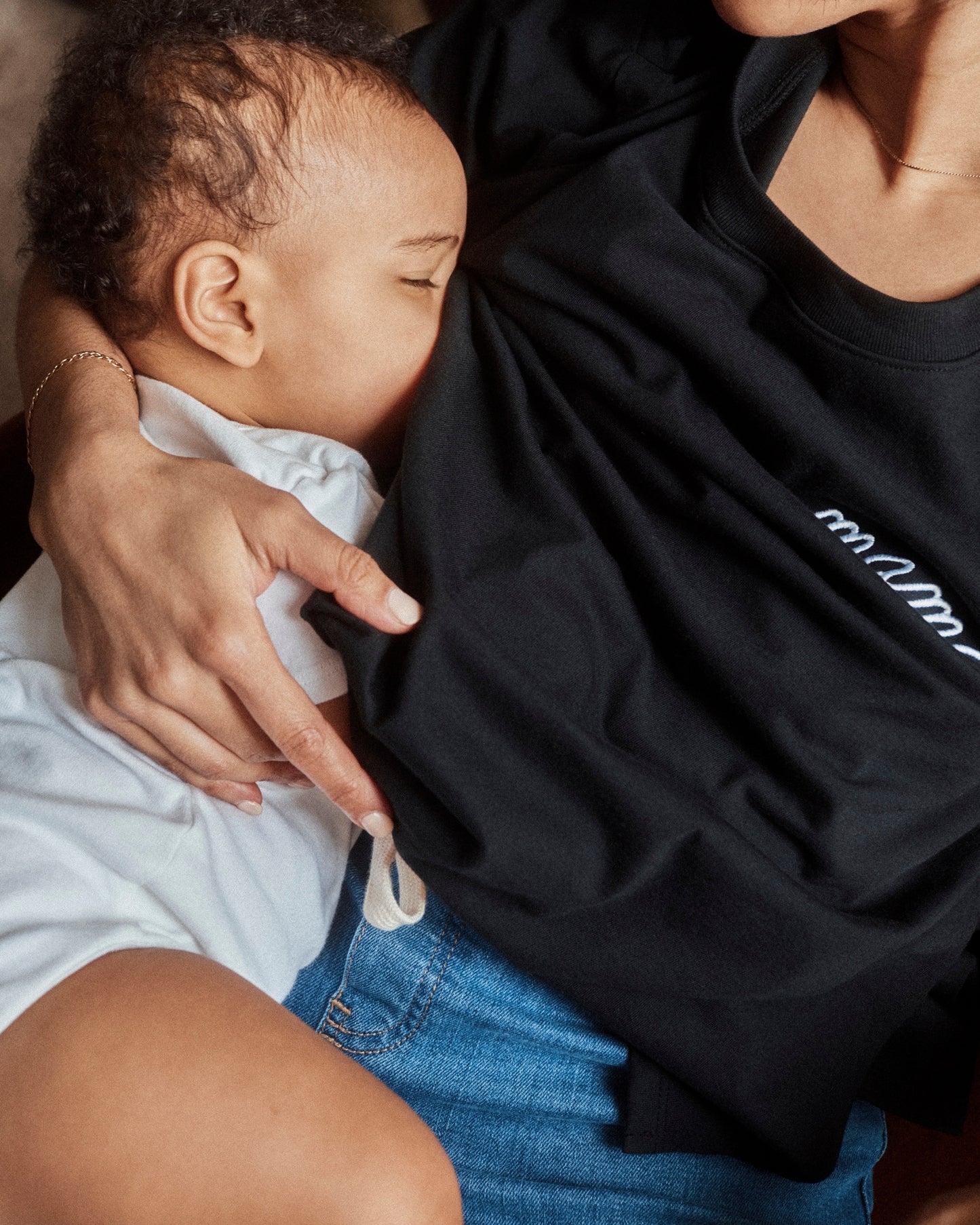 Woman is reclined and breastfeeding baby while wearing Classic Cotton Nursing T-shirt in Black - Embroidered, with the white-threaded word, mama.