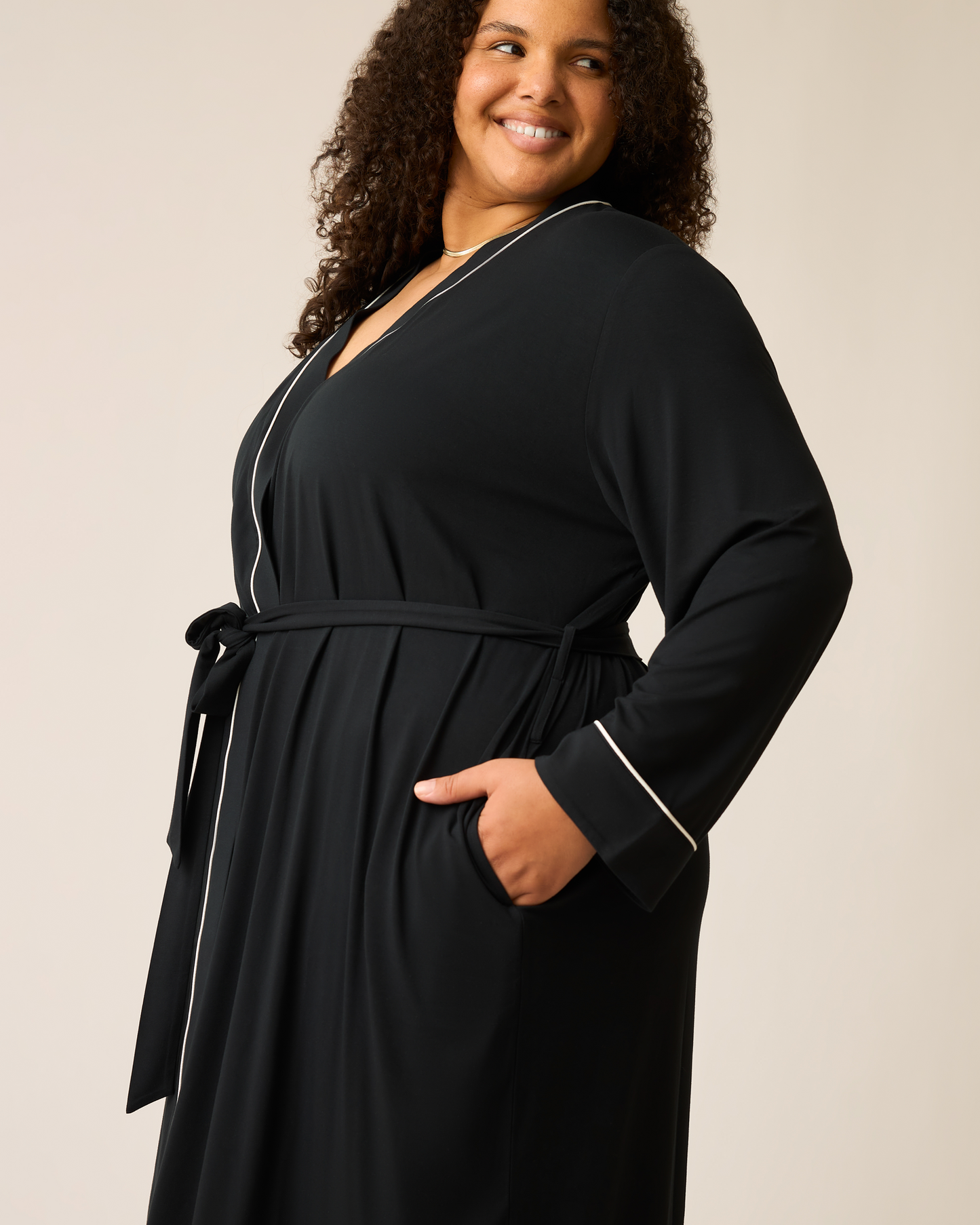 Woman wearing the Clea Bamboo Robe in black with white trim and hand in pocket on a beige background