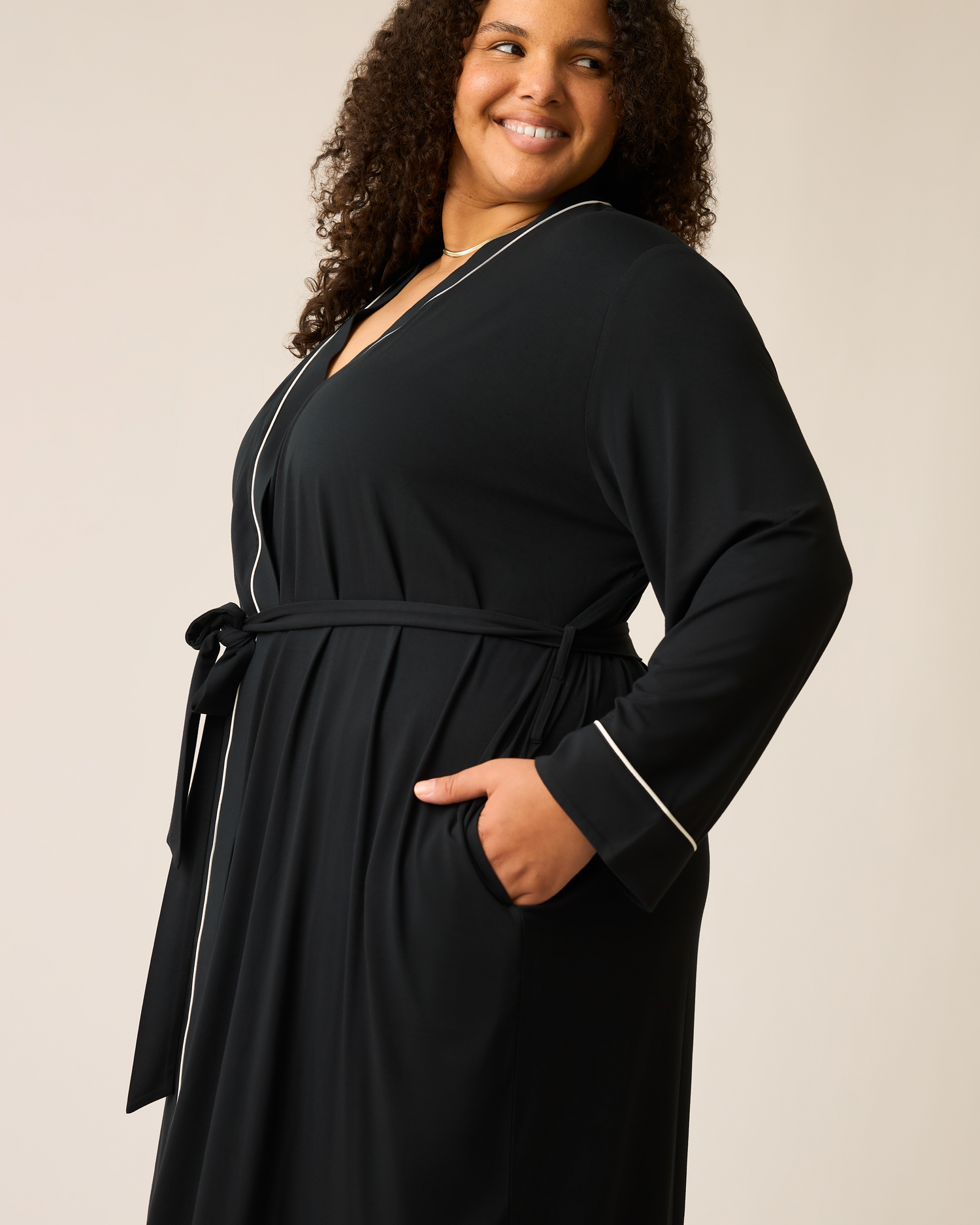 Woman wearing the Clea Bamboo Robe in black with white trim and hand in pocket on a beige background