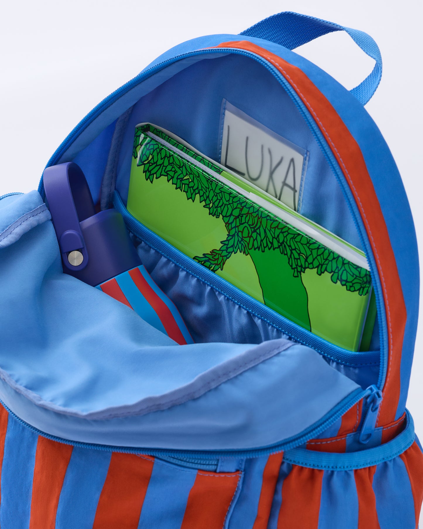 Inside view of the Cool Kid Backpack with name tag, pocket, book and Water Bottle in Blue & Rust Stripe, on a beige background.
