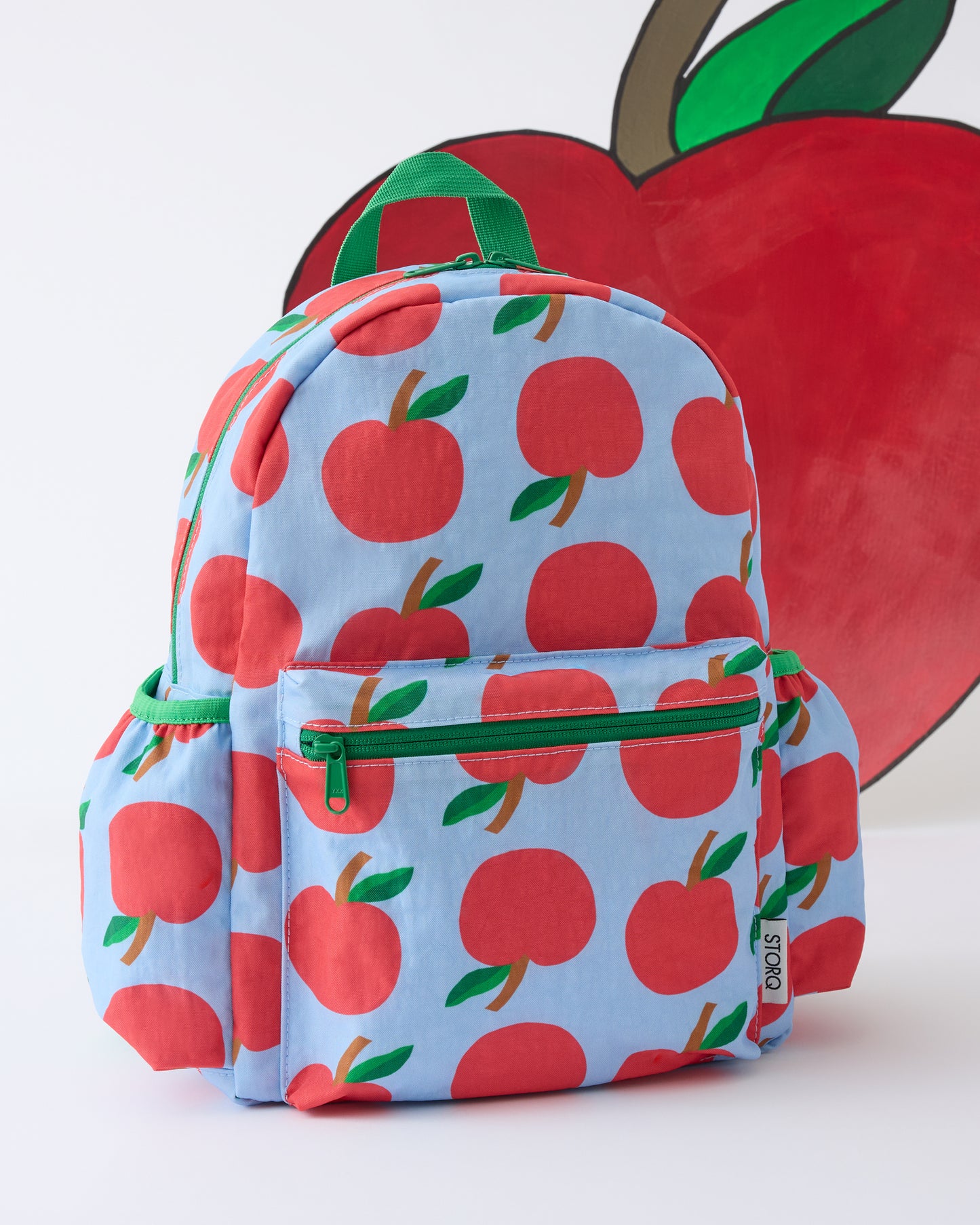 Back view of the Cool Kid Backpack in Blue Apple on a beige background with a giant painted apple behind it.