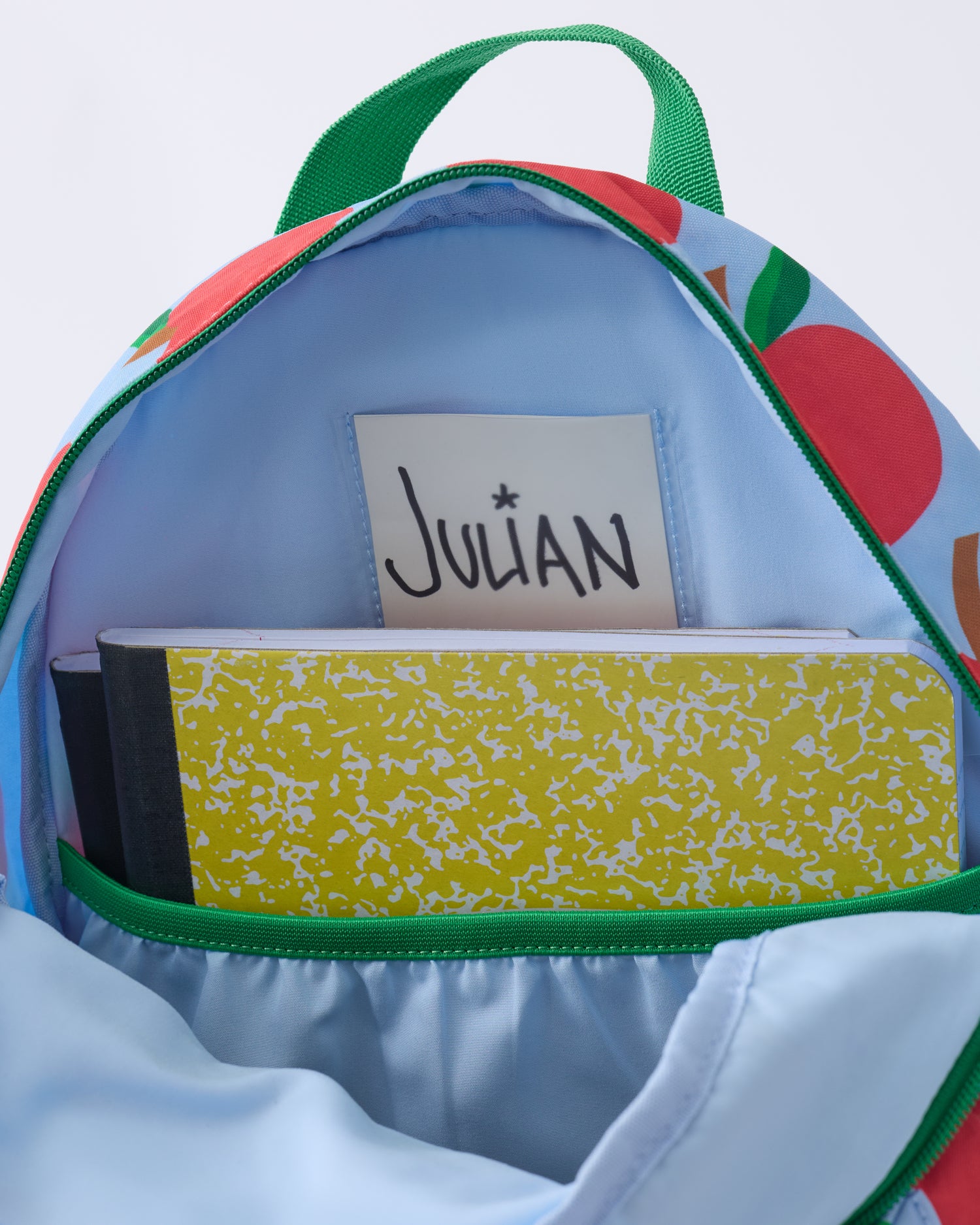 Inside view of the Cool Kid Backpack in Blue Apple with name tag, pocket and notebooks inside, on a beige background.