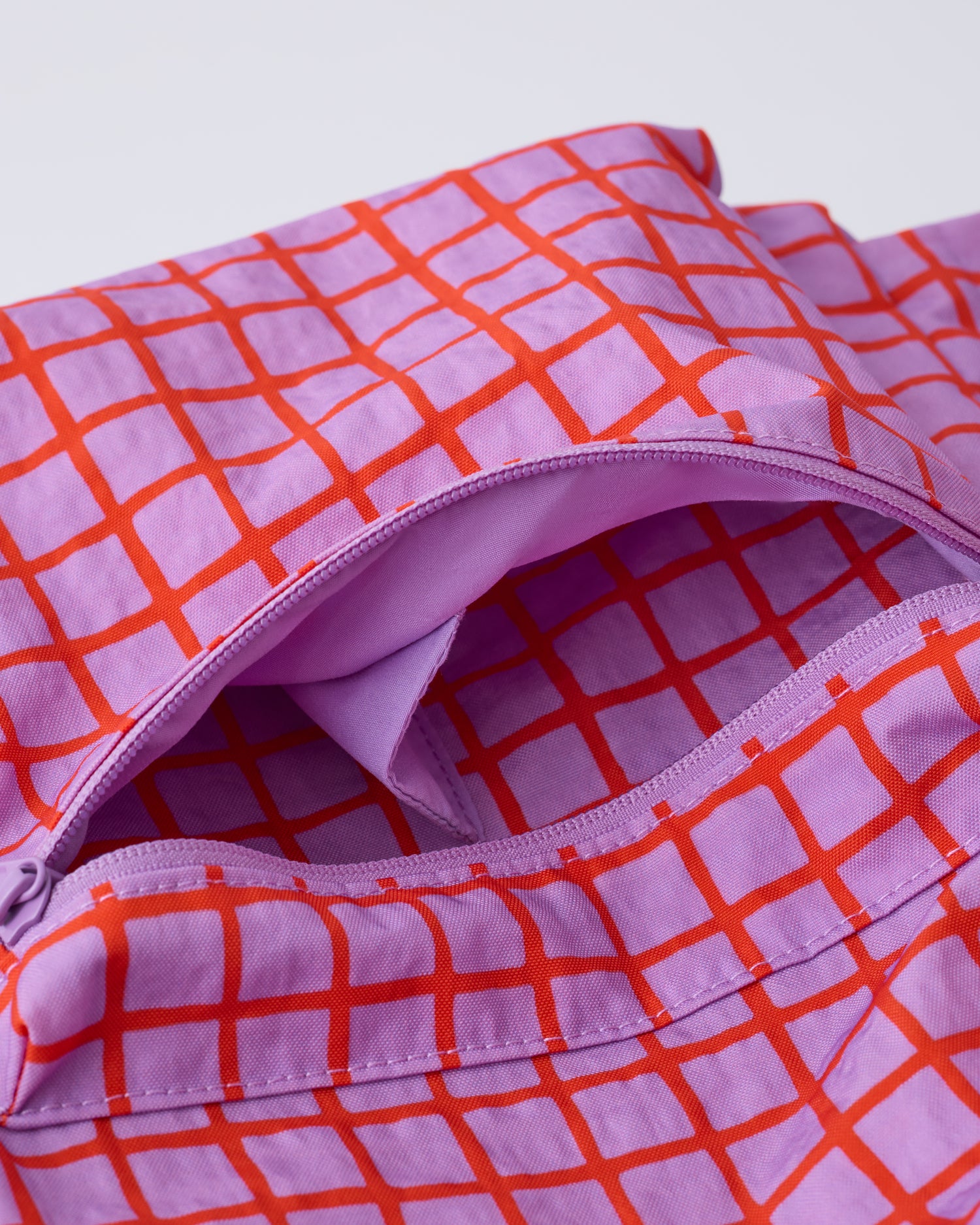 Up close view of the front pocket zipper of the Cool Kid Backpack in Orchid Grid on a beige background.