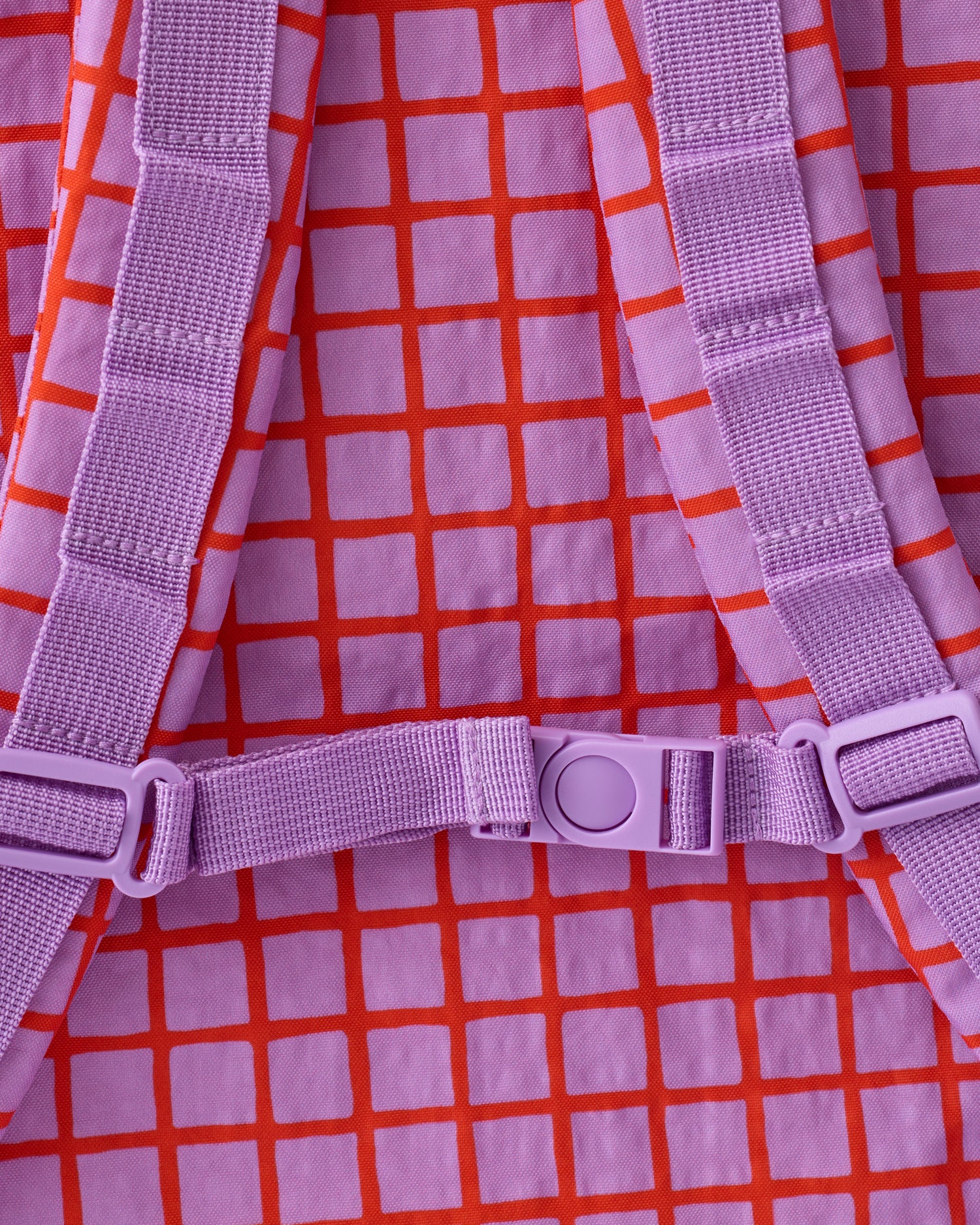 Close up of shoulder strap clip of the Cool Kid Backpack in Orchid Grid on a beige background.
