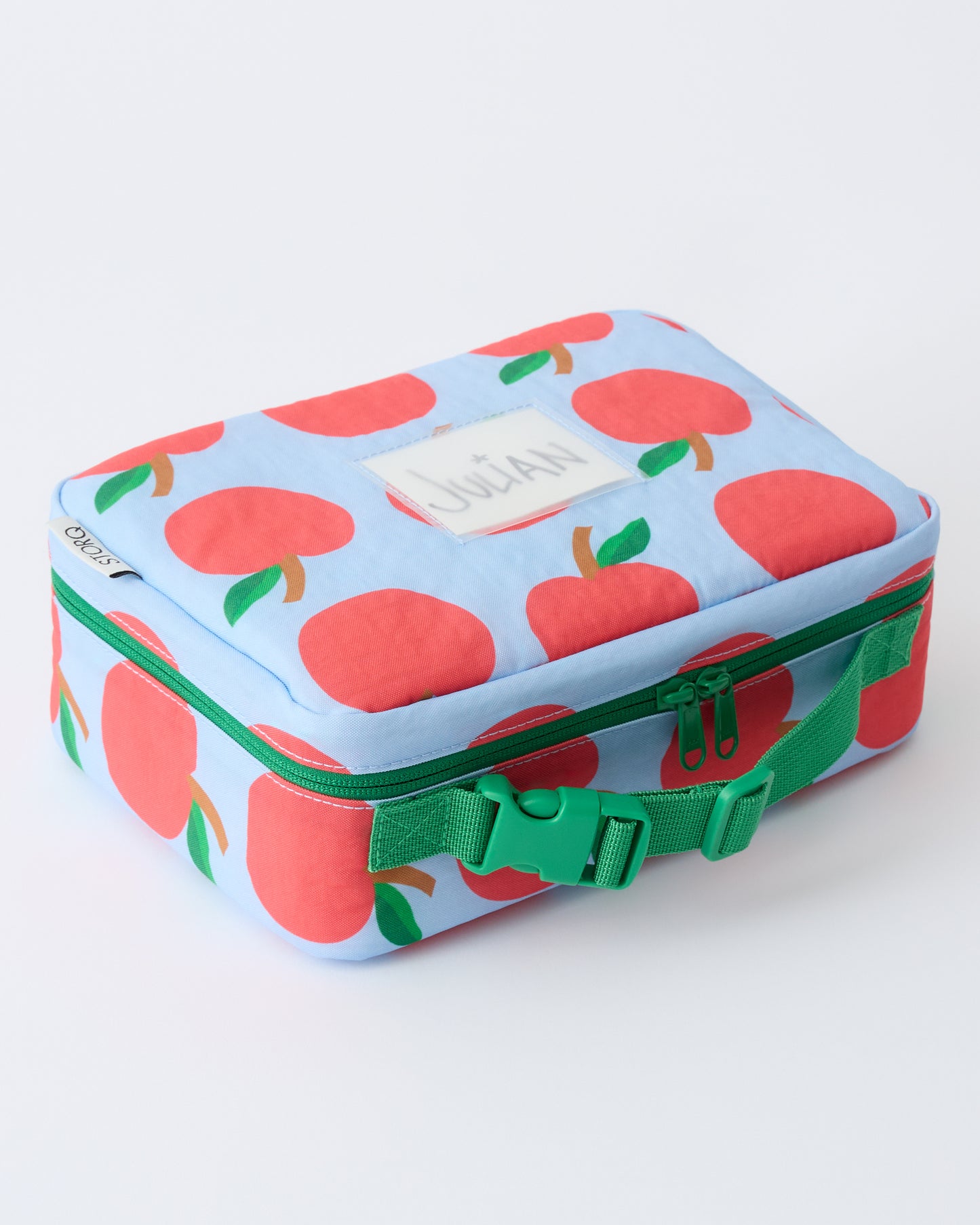 Angled view of Cool Kid Lunch Box in Blue Apple with name tag that reads Julian, on a beige background.