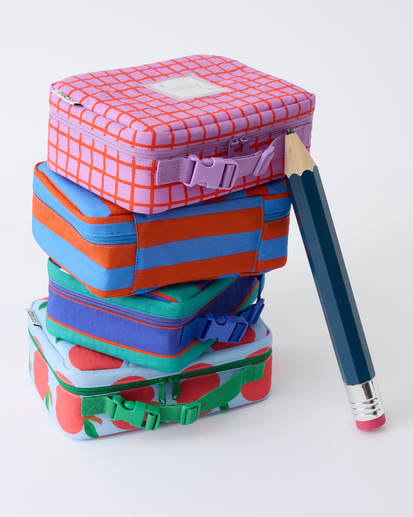 Stack of lunch boxes in order top to bottom: Cool Kid Lunch Box in Orchid Grid, Cool Kid Lunch Box in Blue & Rust Stripe, Cool Kid Lunch Box in Navy & Green Stripe, Cool Kid Lunch Box in Blue Apple, with giant pencil leaning against it, on a beige background.