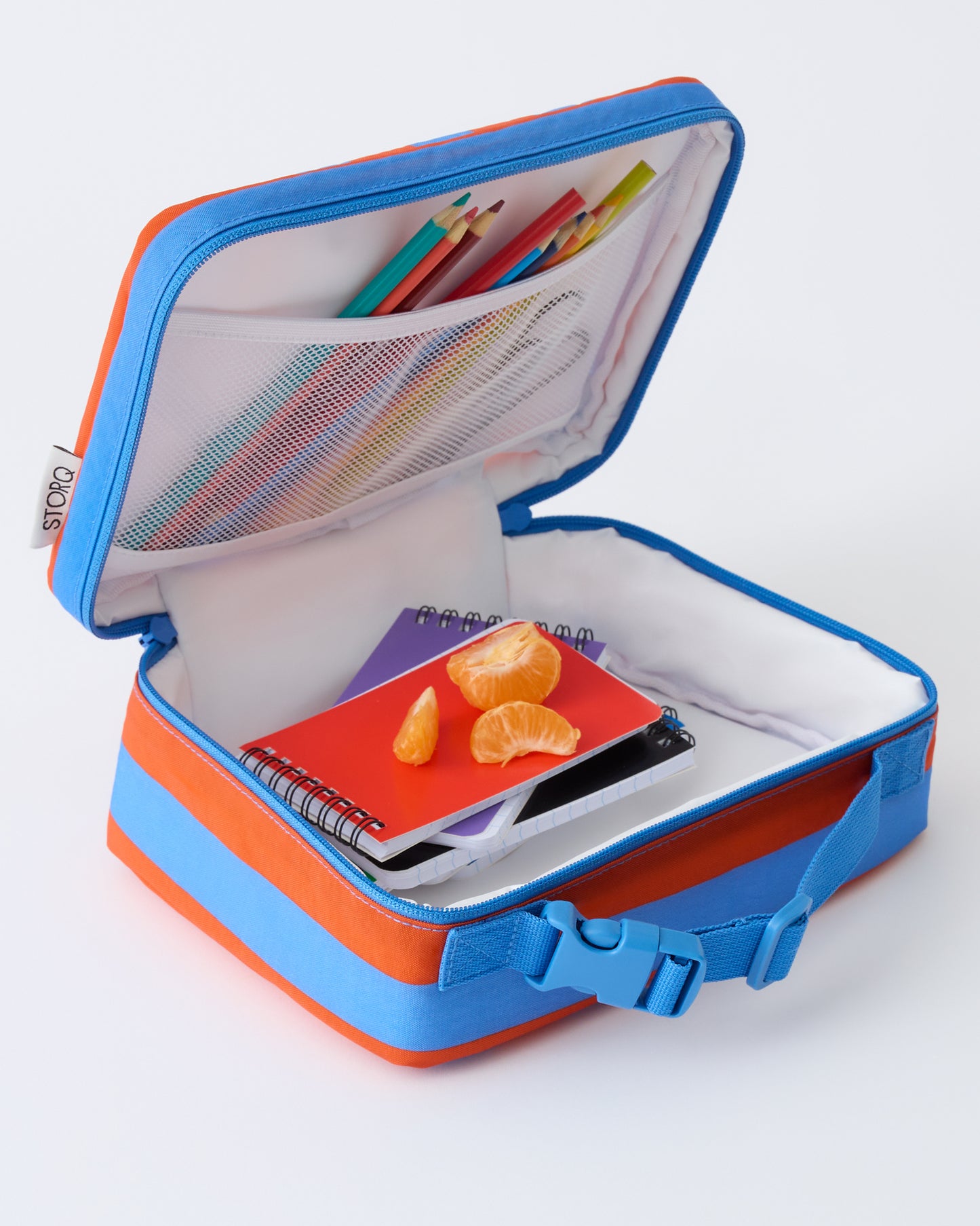 Cool Kid Lunch Box in Blue & Rust Stripe is open with pencils, notebooks and orange slices inside, on a beige background.