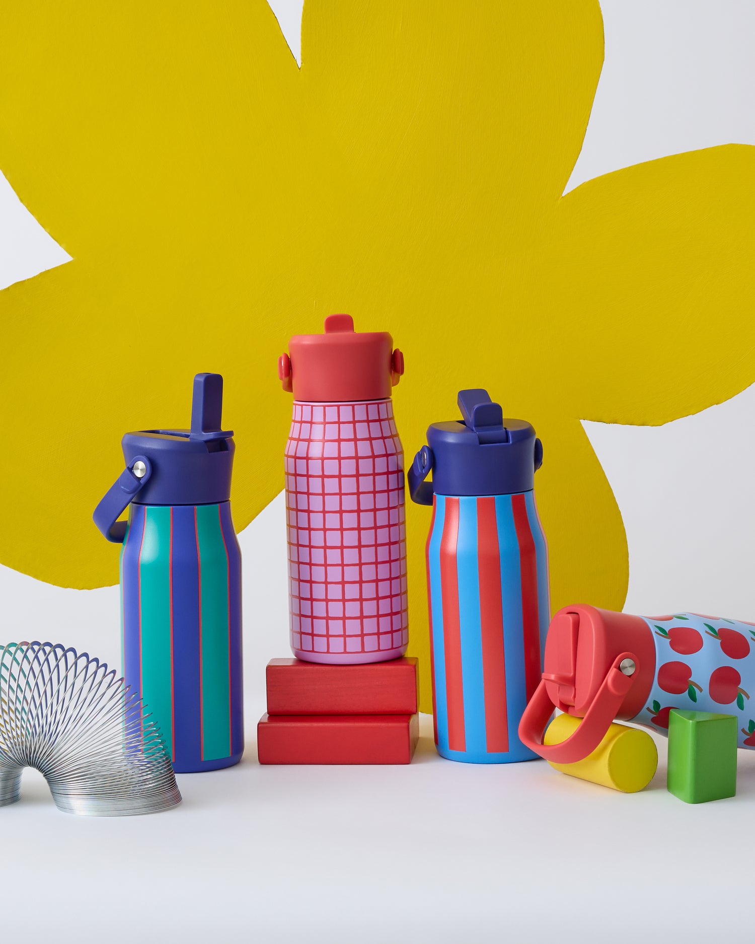 All four Cool Kid Water Bottles, in Orchid Grid, Blue Apple, Blue & Rust Stripe, and Navy & Green Stripe, with toys and a giant playful flower shape behind them, on a beige background.