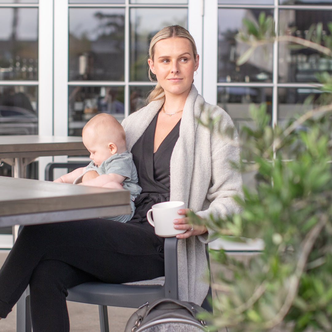 Model sitting at a table with her baby in her lap wearing the Chloe Cardigan Sweater in Grey Heather