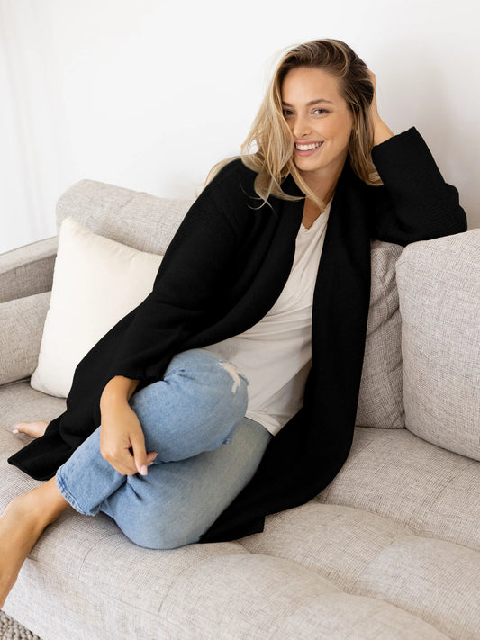 Model wearing the Chloe Cardigan Sweater in Black sitting on a couch. @model_info:Raine is wearing a M/L.