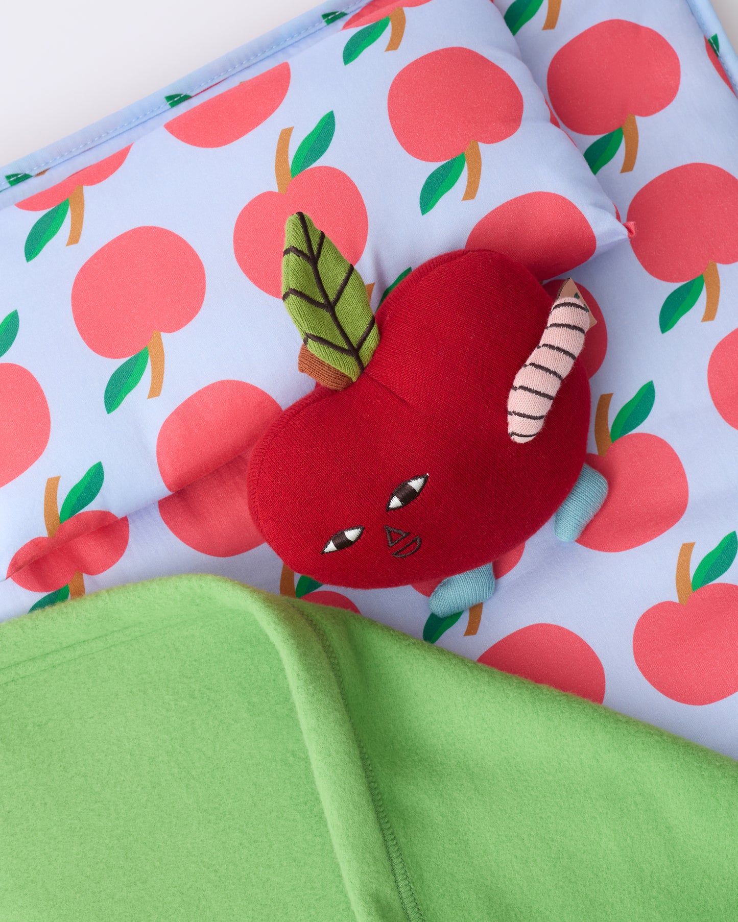 Close up of the pillow and blanket of the Cool Kid Nap Mat in Blue Apple, with small apple lovey leaning on pillow, on a beige background.