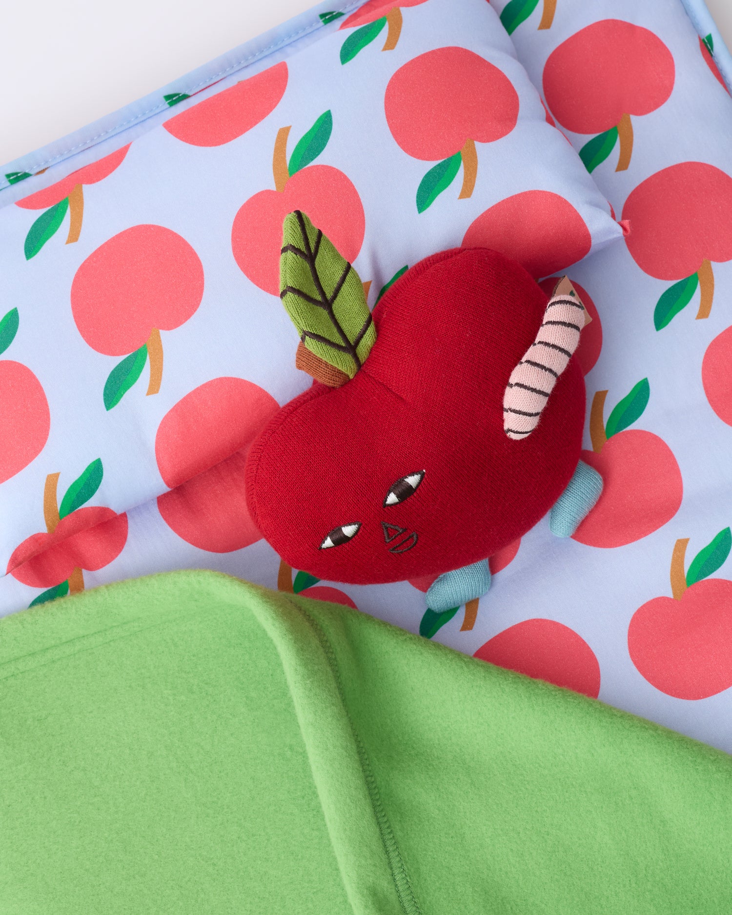 Close up of the pillow and blanket of the Cool Kid Nap Mat in Blue Apple, with small apple lovey leaning on pillow, on a beige background.