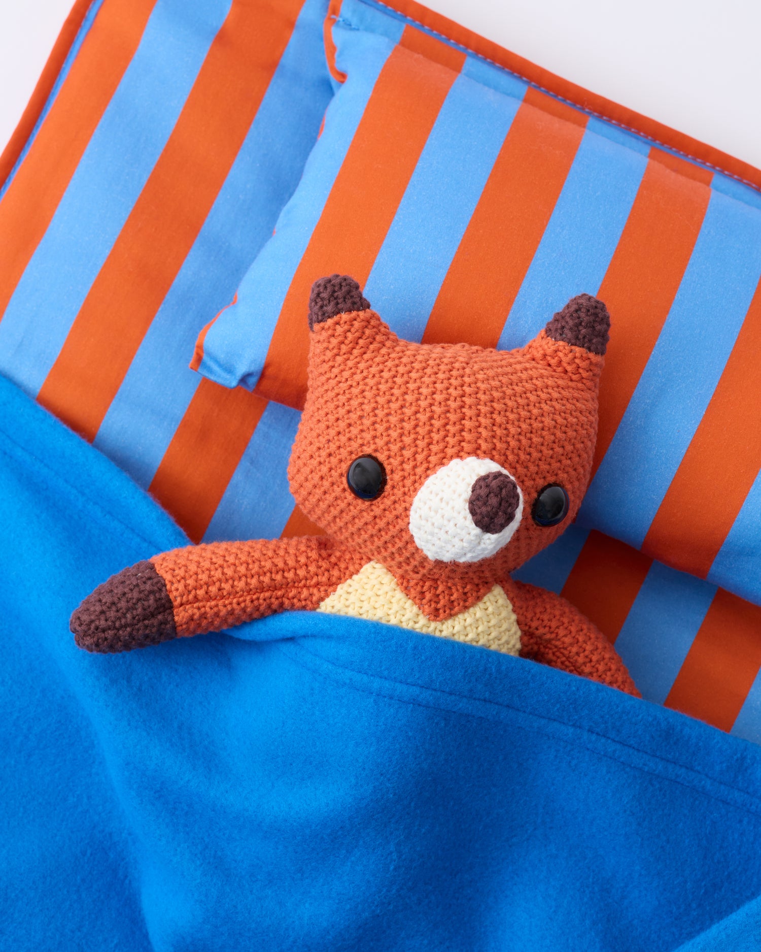 Close up image of the pillow and blanket of the Cool Kid Nap Mat in Blue & Rust Stripe with a little fox lovely resting on the pillow, on a beige background.