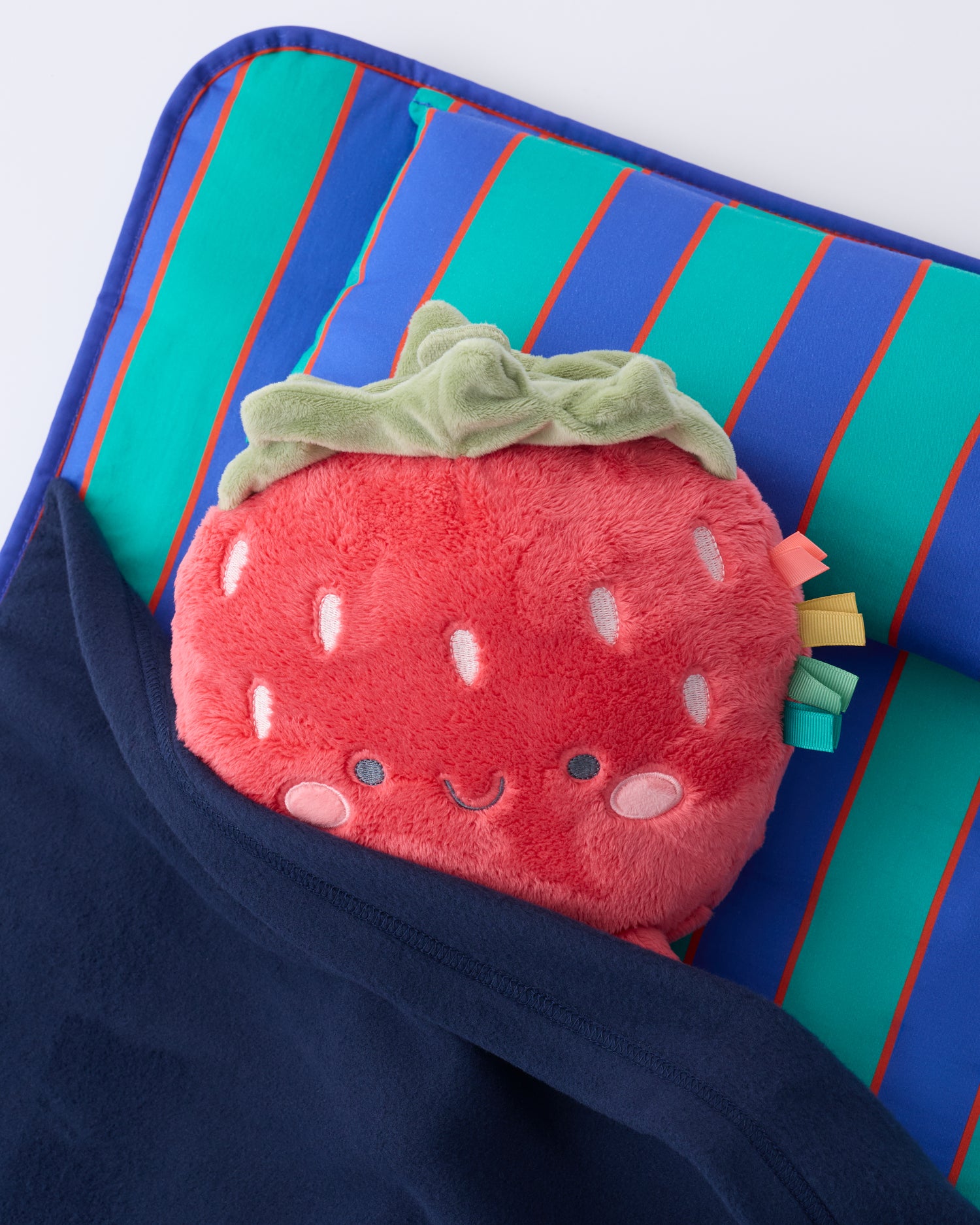 Close up of the pillow and blanket of the Cool Kid Nap Mat in Navy & Green Stripe with a strawberry lovey peeking out of the blanket on a beige background.
