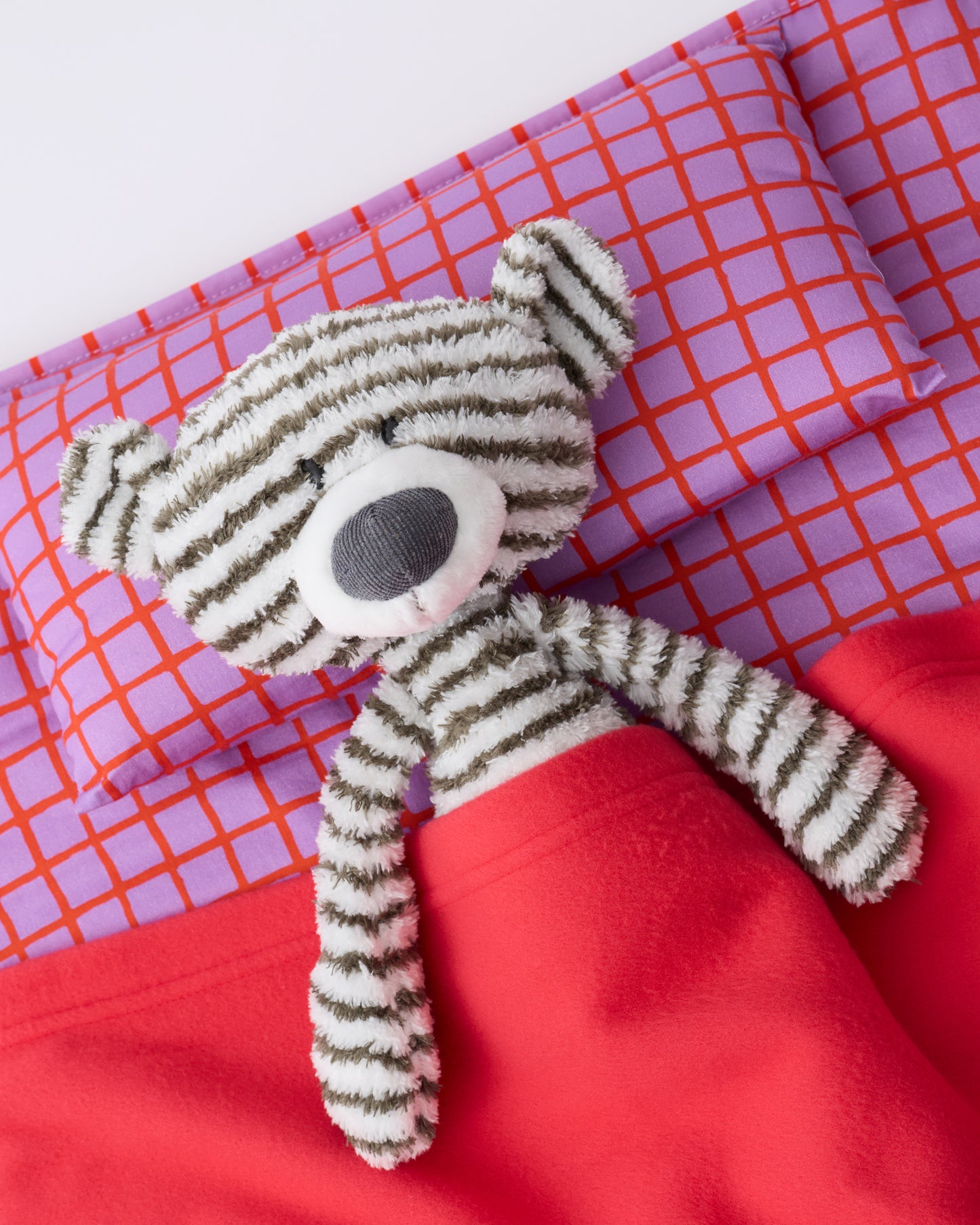 Close up of the pillow and blanket of the Cool Kid Nap Mat in Orchid Grid, with a little striped bear lovey laying under the blanket, on a beige background.