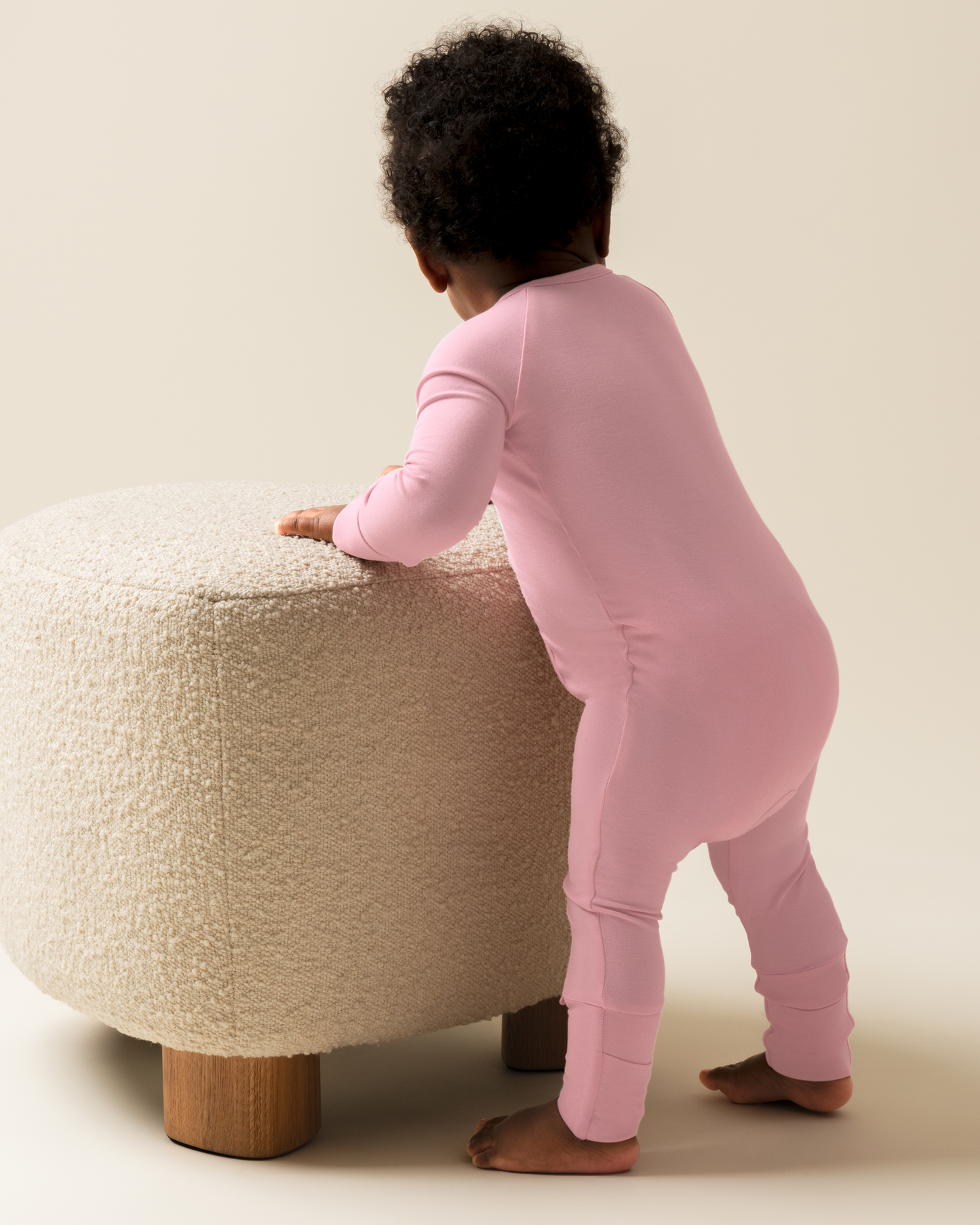 Child in Bamboo Baby Pajamas in Light Mauve standing next to a textured stool on a plain background
