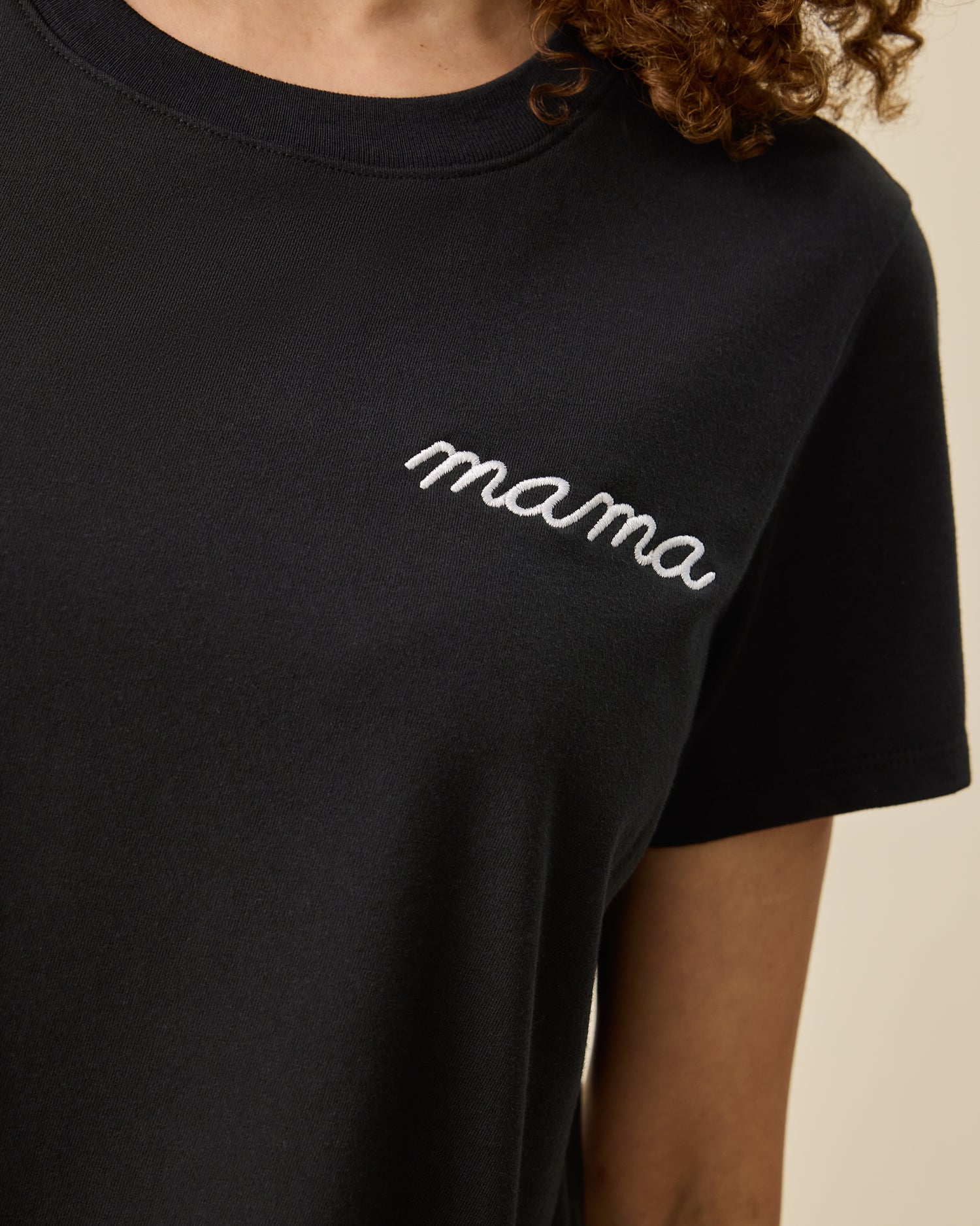 Close up of Classic Cotton Nursing T-shirt in Black - Embroidered, with the white-threaded word, mama, on a beige background.
