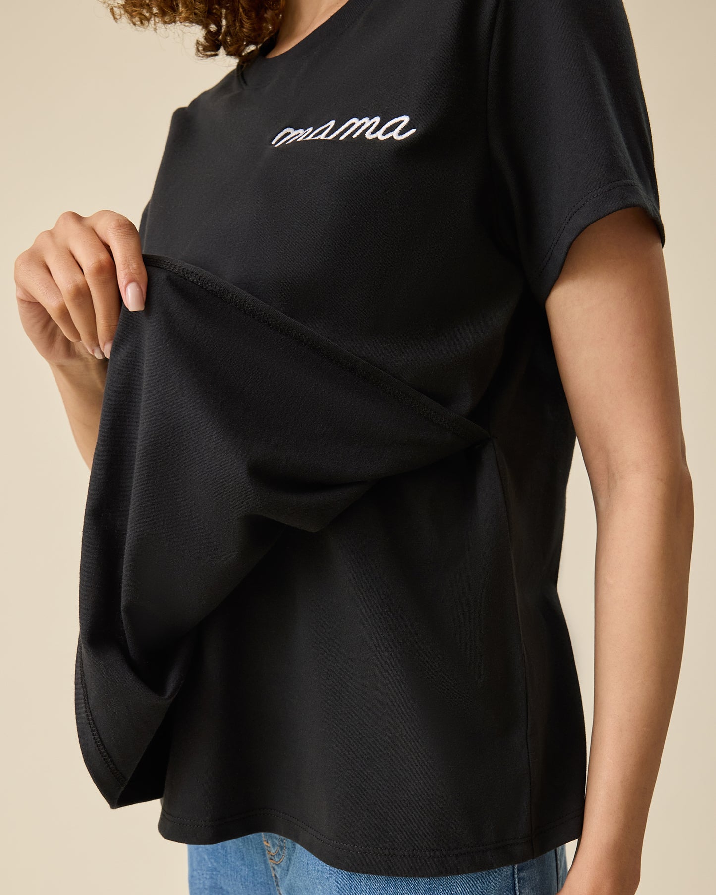 Model is flipping up the front panel of the Classic Cotton Nursing T-shirt in Black - Embroidered, with the white-threaded word, mama, on a beige background.