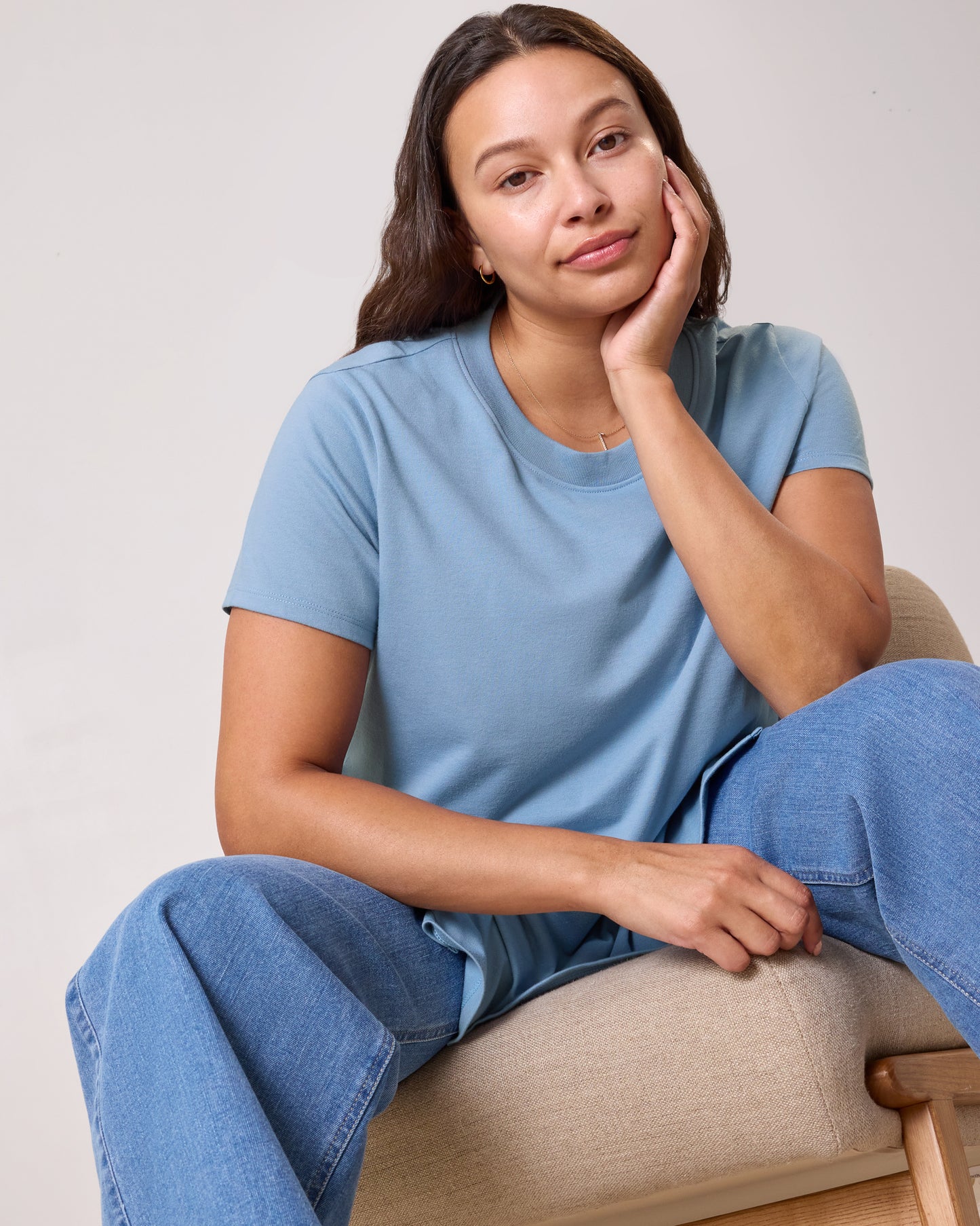 Model wearing Classic Cotton Nursing T-shirt in Vintage Blue on a neutral background, sitting on a lounging chair and in a relaxed pose leaning forward