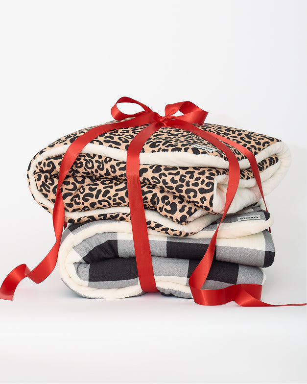 Cozy Stroller Blanket in Leopard and Cozy Stroller Blanket in Buffalo Plaid stacked and tied together with a big red bow, on an off white background.