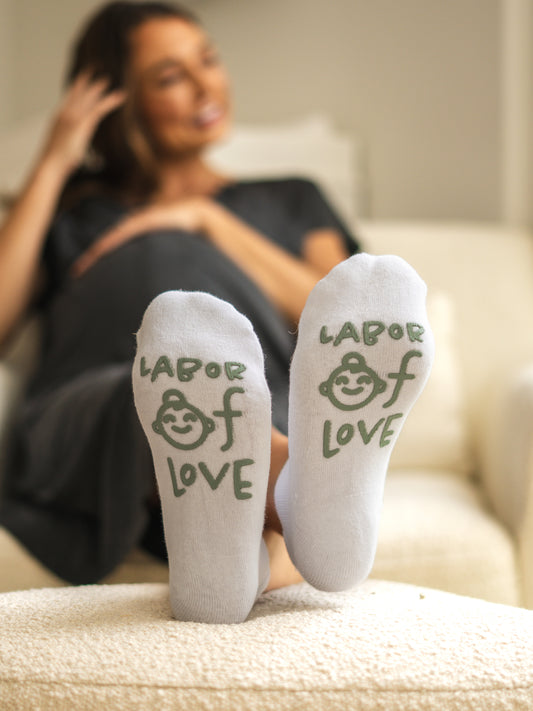 Model sitting on chair with feet on ottoman wearing Labor of Love Labor & Delivery Socks in sage