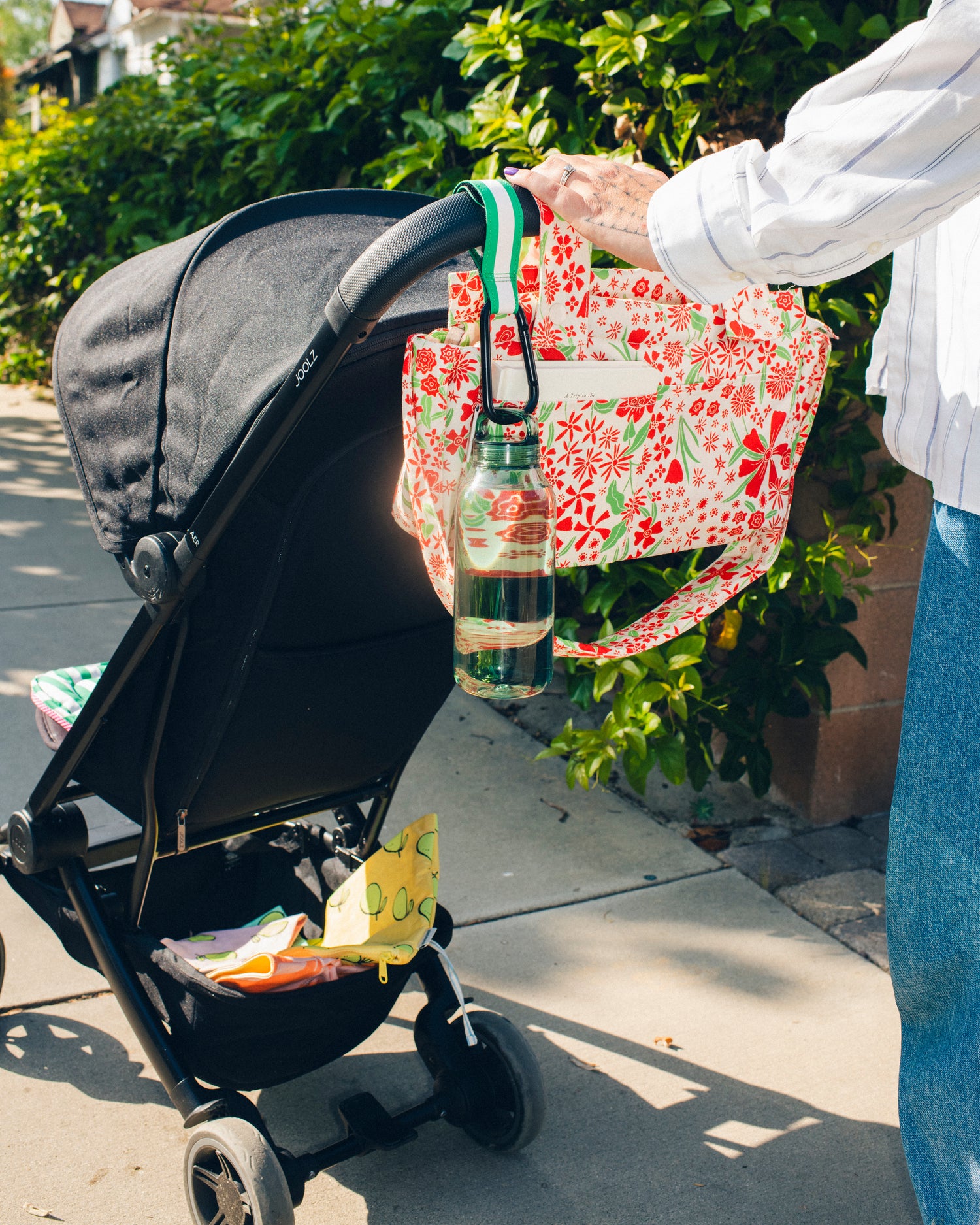Woman is pushing stroller down sidewalk with Stroller Party™ Caddy Bag in Superbloom paired with Stroller Party™ Clip in Green Stripe to hold water bottle. Zipper Pouch Three-Piece Set in Pomelo are sitting in bottom carrier, ready to help organize throughout the day.