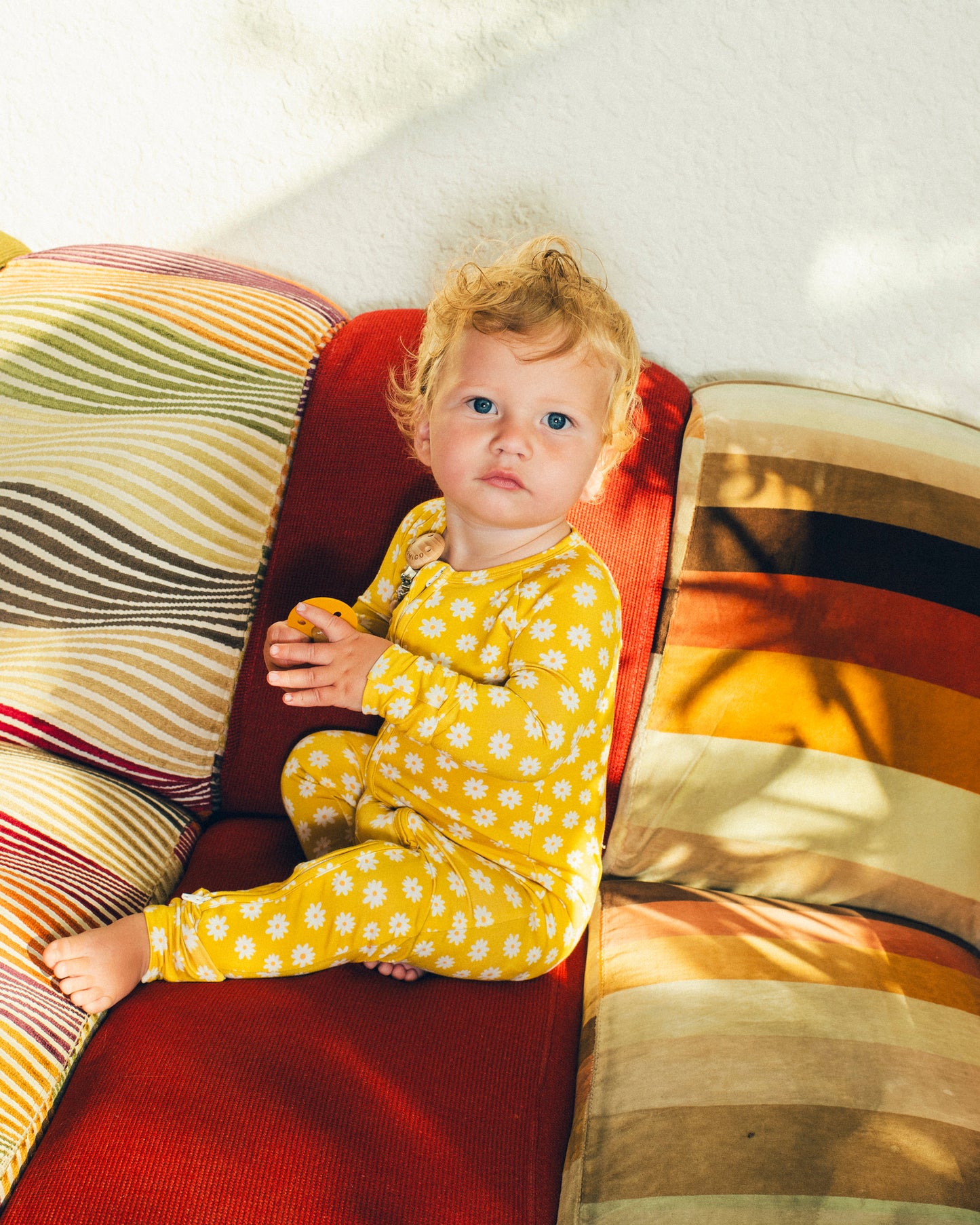 Baby is sitting on sunny outdoor couch in various striped patterns and holding pacifier while wearing Bamboo Baby Pajama in Yellow Daisies.