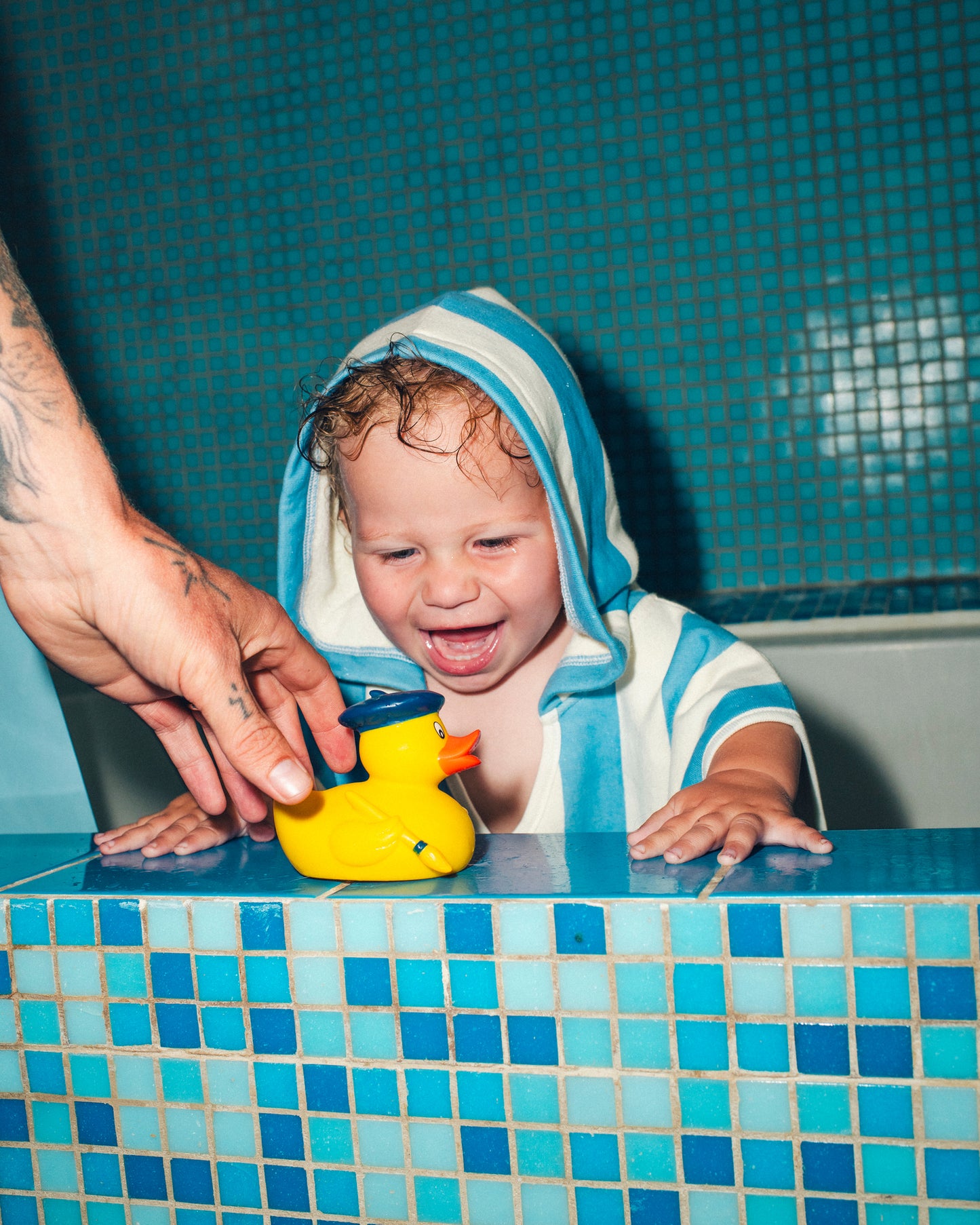 Toddler is standing in blue tiled bathtub, laughing at rubber ducky, while wearing Organic Cotton Toddler Poncho Towel in Blue Wide Stripe.